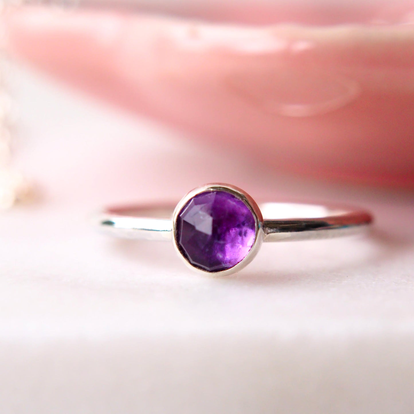 Amethyst ring made from Sterling SIlver and a round 5mm facet cut cabochon Amethyst. Pictured on a white background with a pink trinket dish in the background. Made by Maram Jewellery