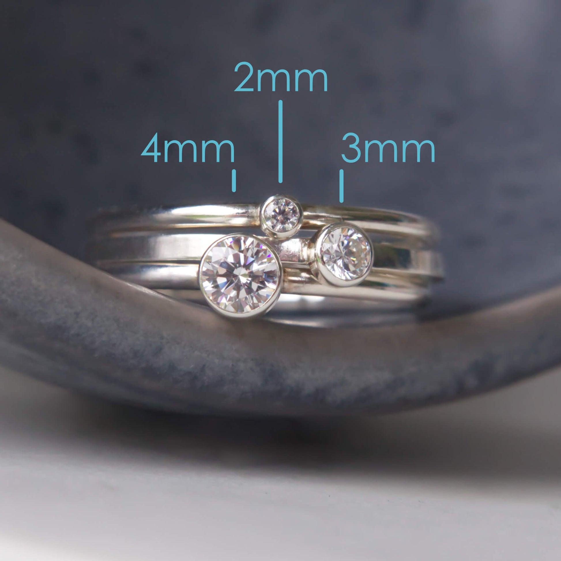 Trio of Diamond Silver Cubic Zirconia Solitaire rings with round stones. Each of the rings have a different sized gem in a 2mm, 3mm and 4mm size, with text to indicate size. Pictured on a grey ceramic background. Rings handmade by maram jewellery in Scotland UK