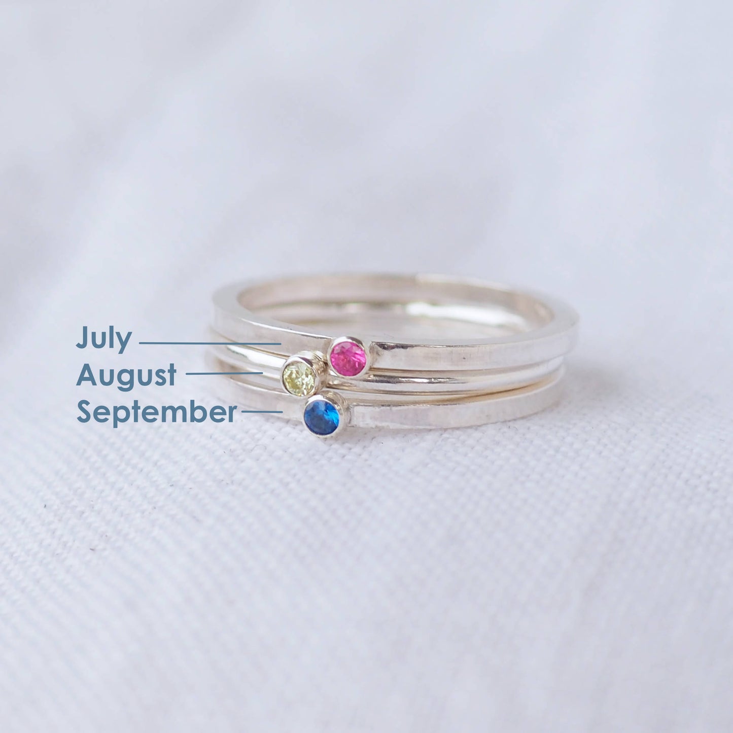 Three silver rings showing April, May and June Birthstones. The rings are simple silver bands with a miniature birthstone on the band in clear, green and pink cubic zirconia. Handmade in Edinburgh by maram jewellery