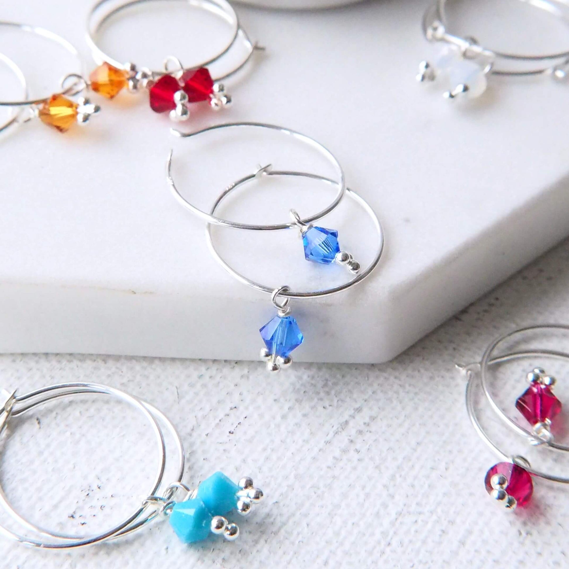 Group shop of maram jewellery birthstone crystal earrings with blue sapphire crystal version at front. the earrings are a thin sterling silver full hoop, 2 cm diameter with a blue sapphire crystal charm drop for September's birthstone. handmade in Scotland