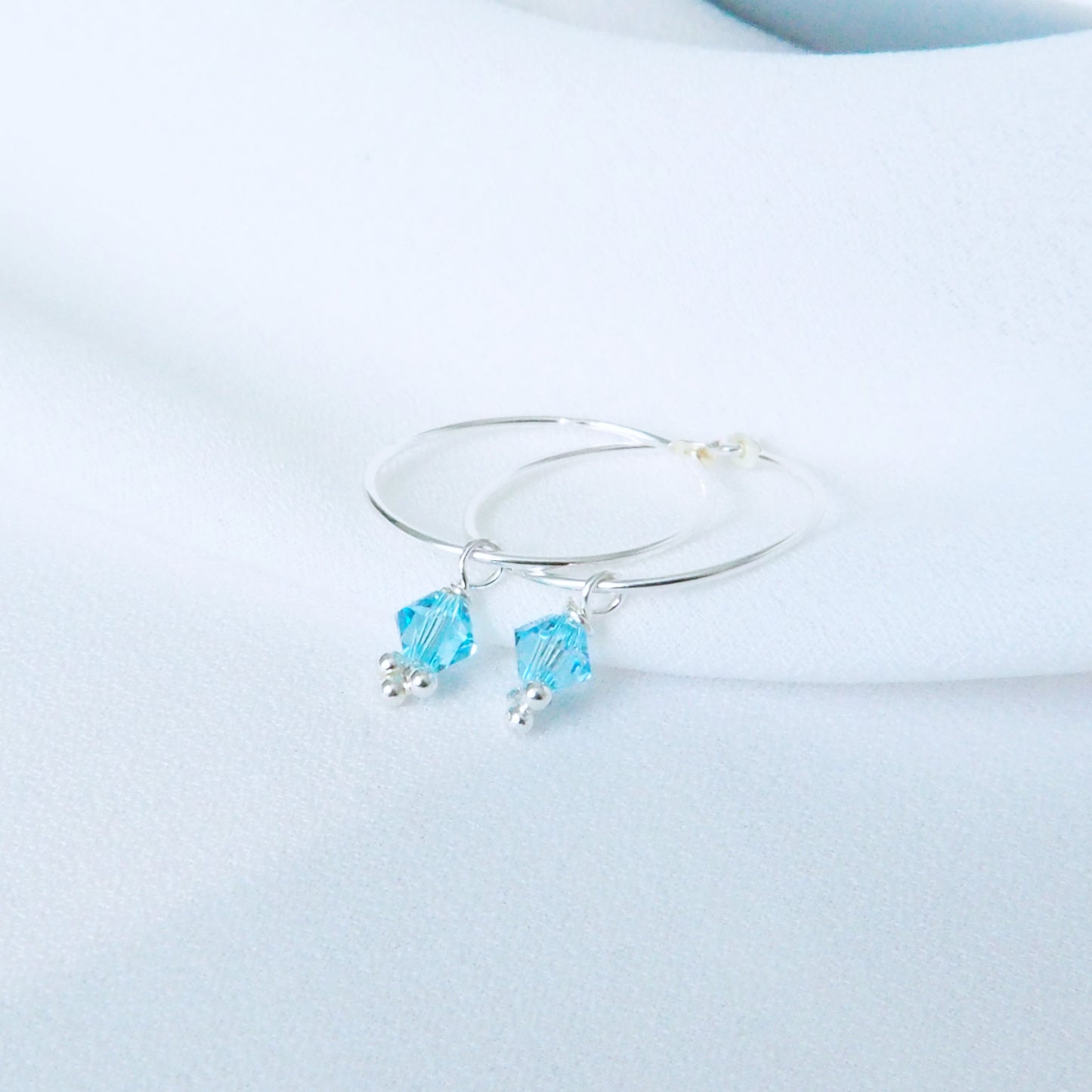 Pale blue aquamarine crystals in a diamond shape dangle from a fine silver hoop earring to create a simple modern birthstone earring. The earrings are pictured on a fine satin white material