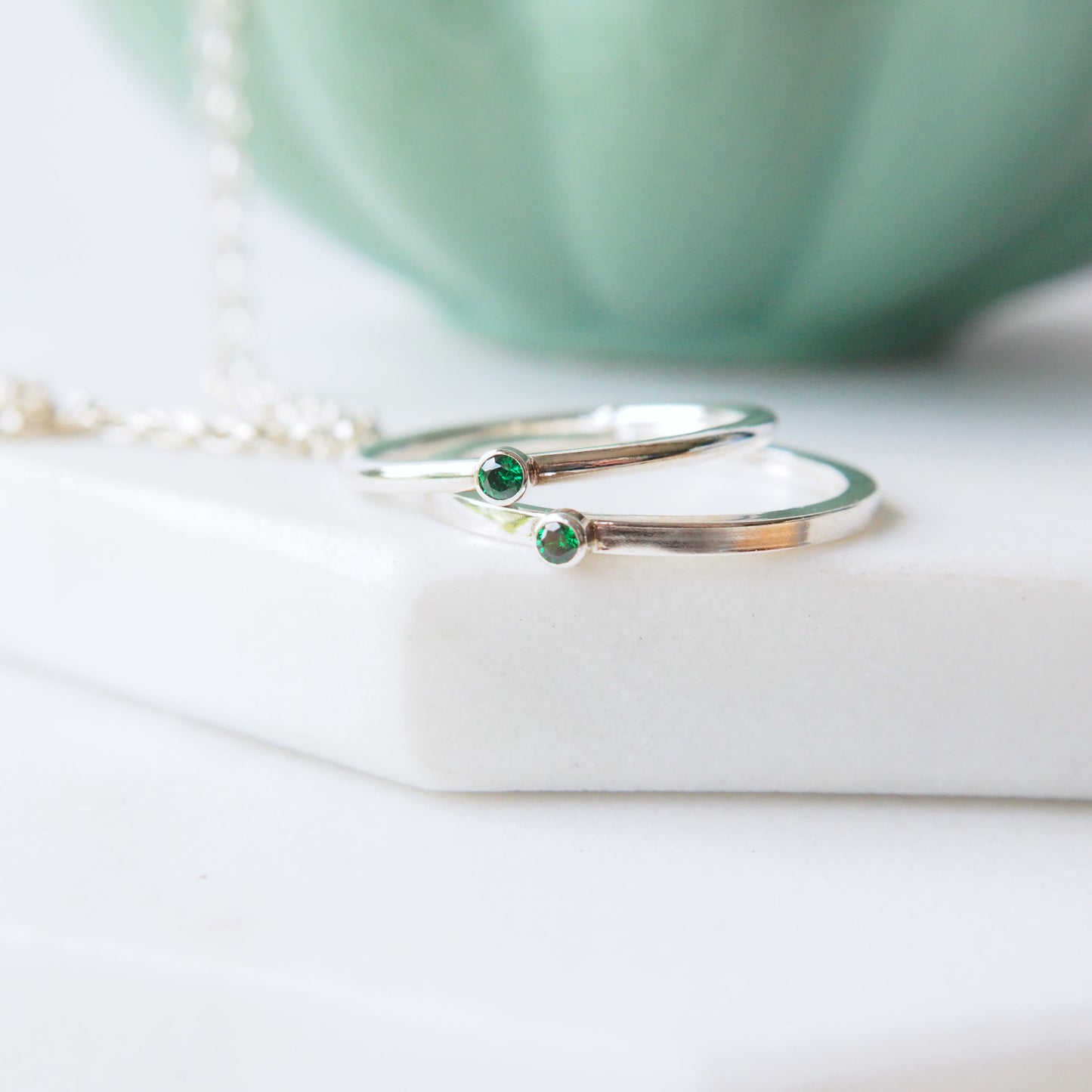 two silver ring with a very small 2mm emerald cubic zirconia. Handmade by maram jewellery. shown on a white marble slab with a green jewellery bowl in the background