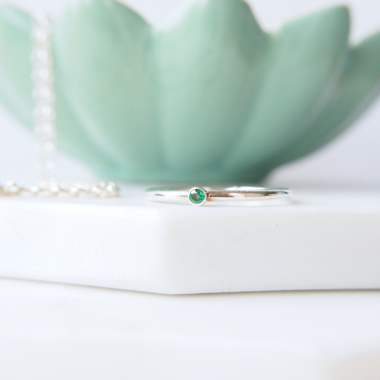 round band silver ring with a very small 2mm emerald cubic zirconia. Handmade by maram jewellery. shown on a white marble slab with a green jewellery bowl in the background, Made in Scotland