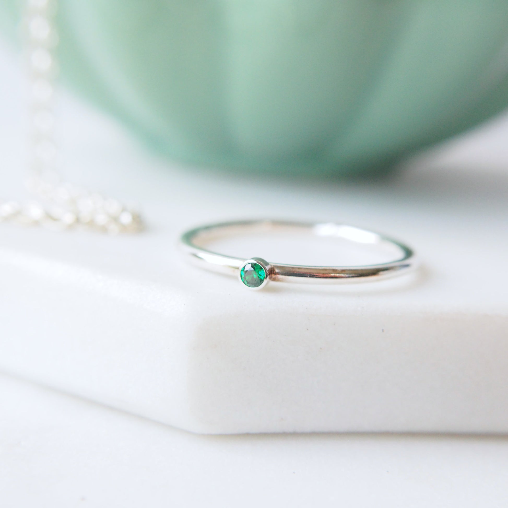 Round band silver ring with a very small 2mm emerald cubic zirconia. Handmade by maram jewellery. shown on a white marble slab with a green jewellery bowl in the background