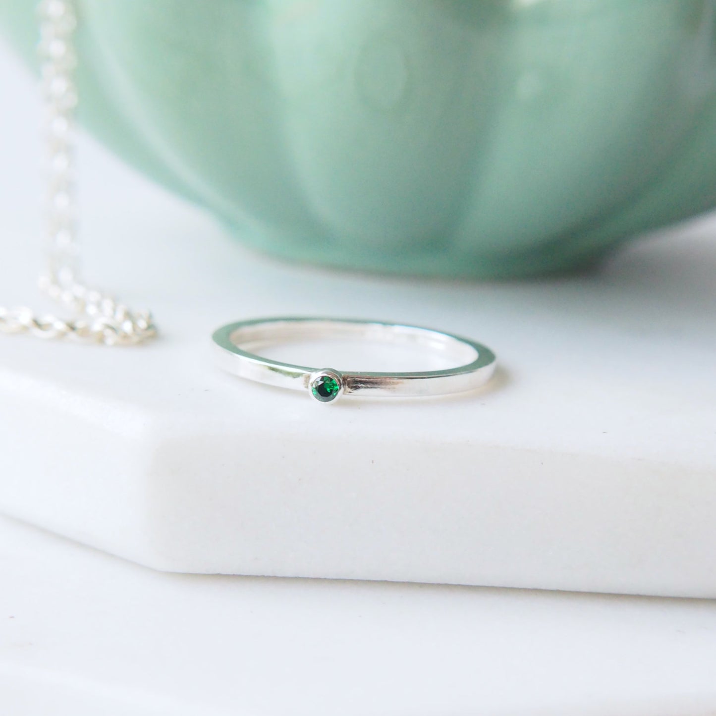 square band silver ring with a very small 2mm emerald cubic zirconia. Handmade by maram jewellery. shown on a white marble slab with a green jewellery bowl in the background