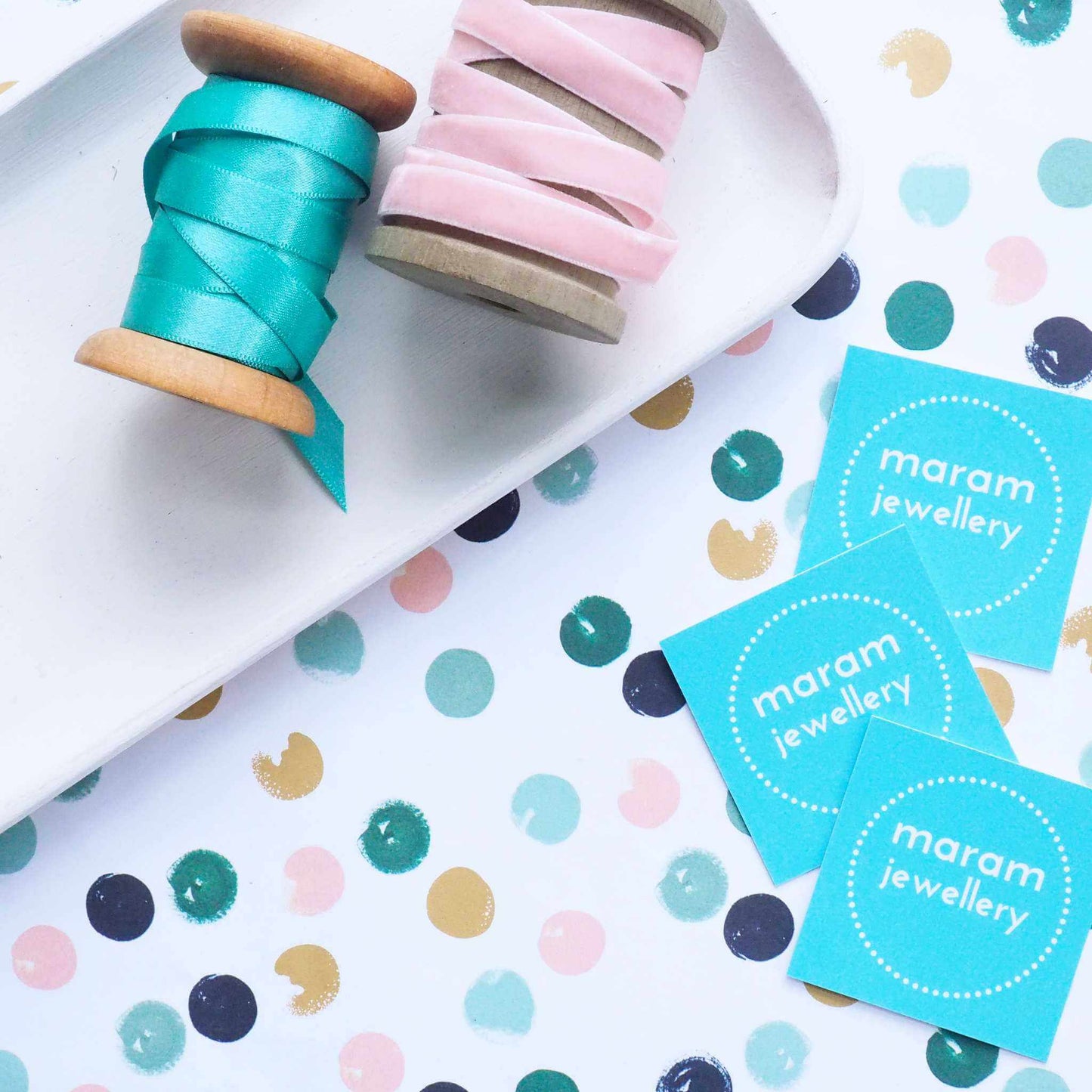 Flatlay style image of maram jewelly gift wrap paper, white with green and pink dots, with a tray alongside it with business cards and ribbons