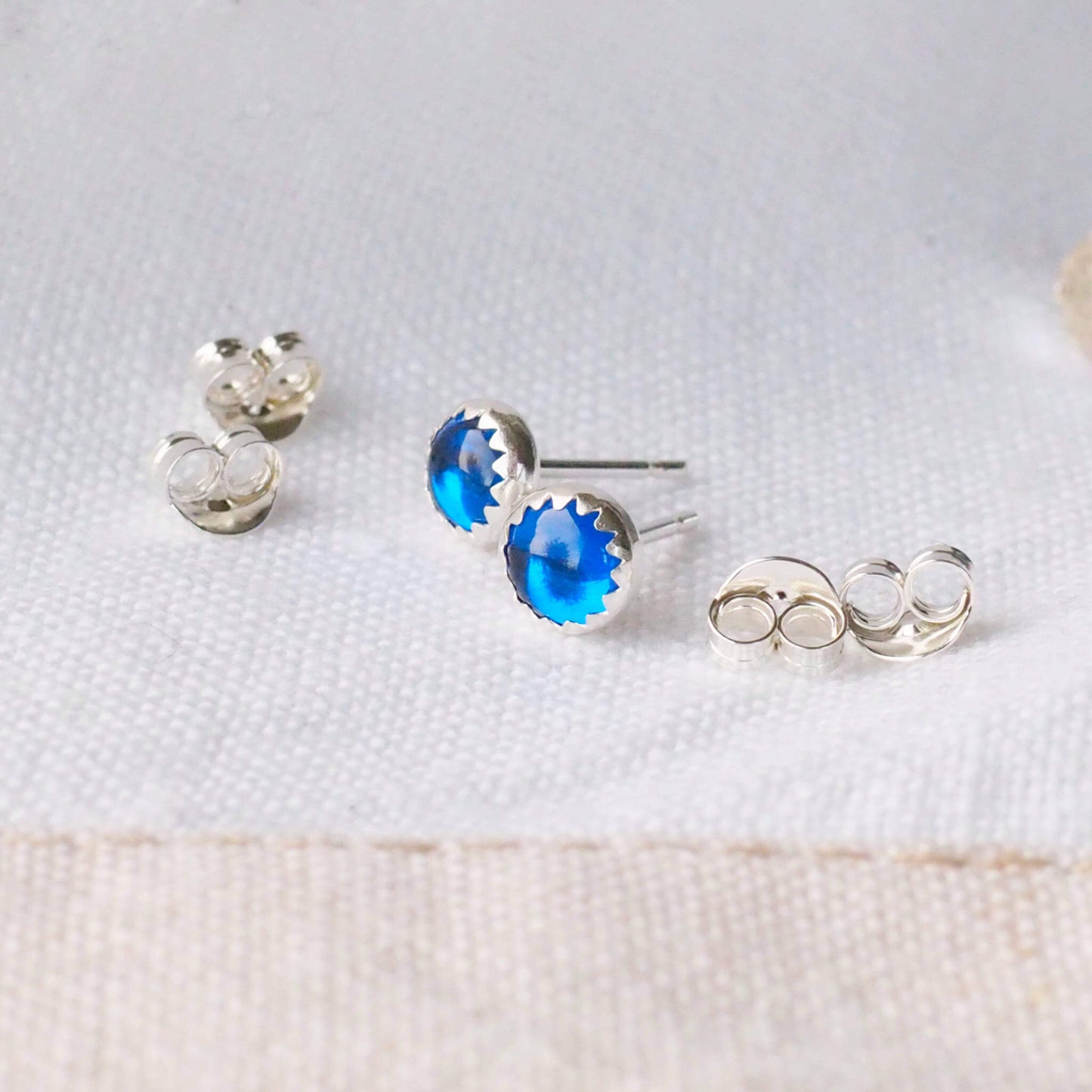 Pair of Sapphire Blue and Silver Gemstone studs with a round cabochon gemstone set into a simple sterling silver mount. the earrings are round measuring 5 mm in diameter and are pictured on a white linen background with earring backs at the side. Handcrafted by maram jewellery in Scotland