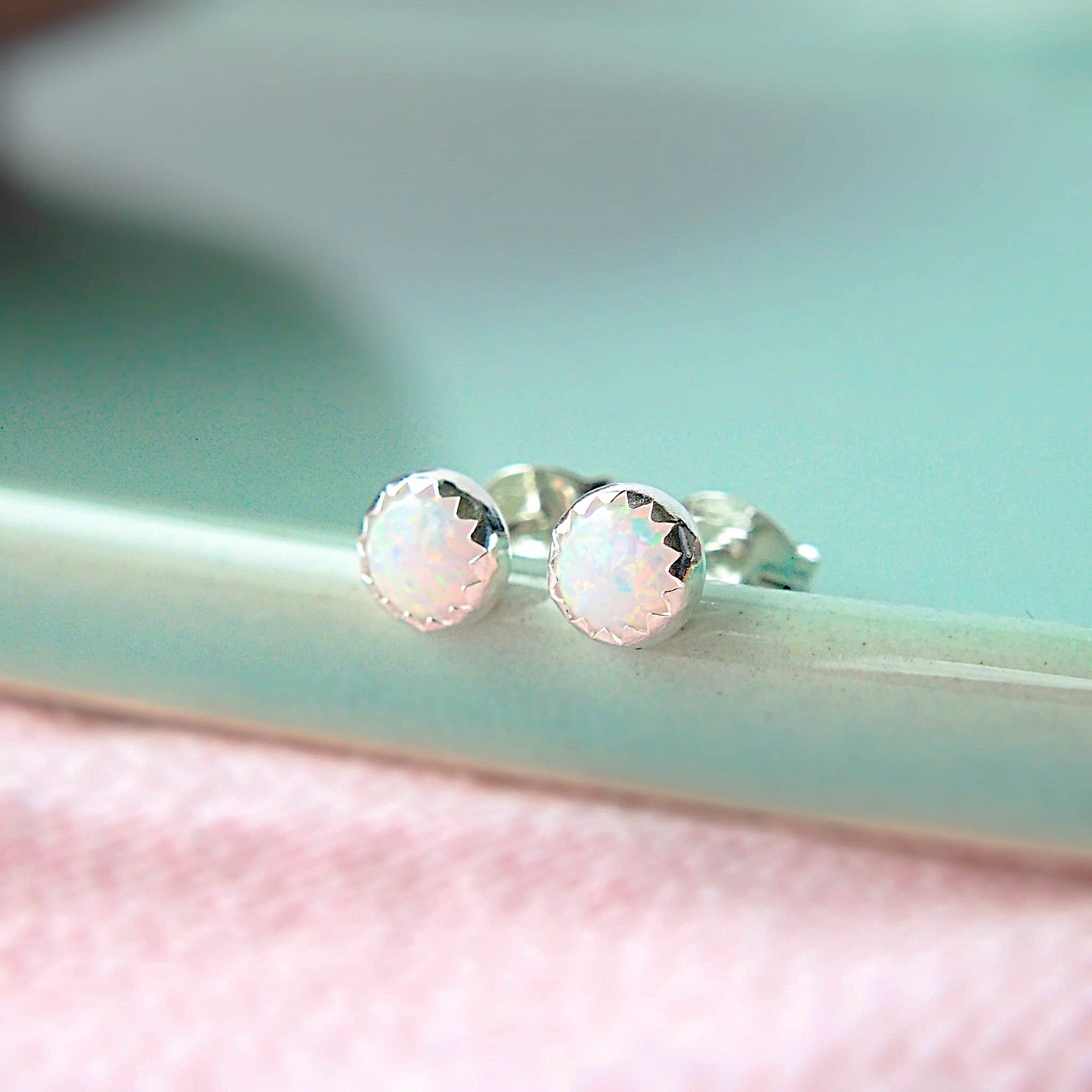 A pair of lab opal gemstones studs sit on the edge of a pale green plate on a pink tablecloth. The earrings are made from 5mm round white lab opals with a zig zag silver edge. Handcrafted by maram jewellery in the UK