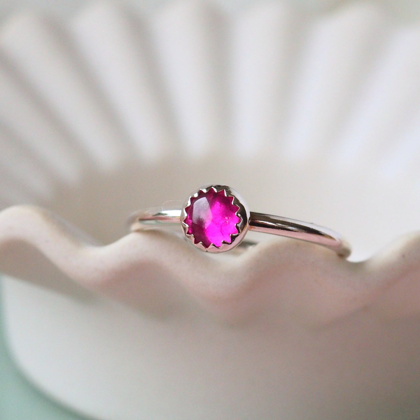 Bright Pink and Silver Gemstone ring in a modern simple style with a round cabochon gemstone. The ring is made from sterling silver and features an unusual zig zag edge where the stone meets the silver. the ring is photographed on a white ceramic bowl with a wavy edge. Handmade by maram jewellery in Scotland