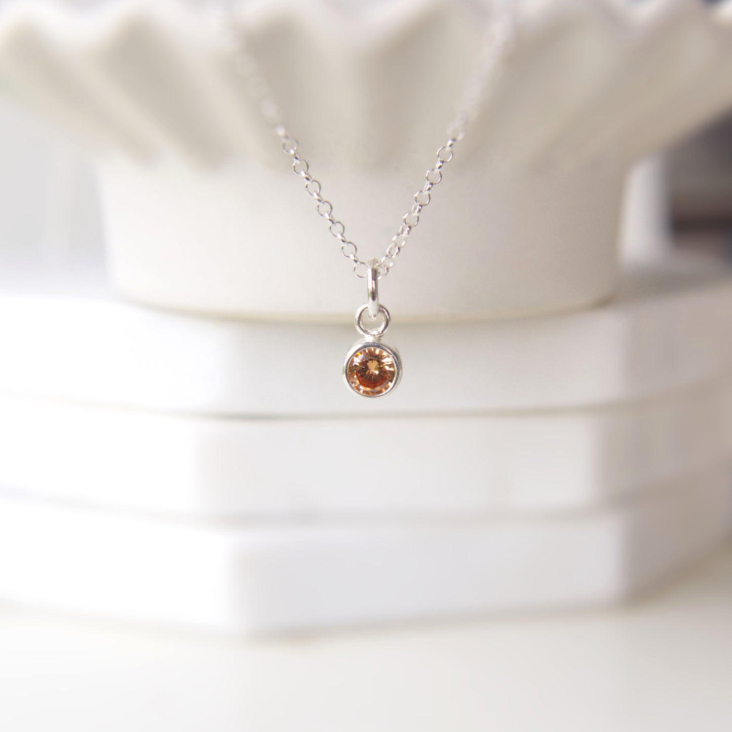 A simple solitaire gemstone pendant in a honey yellow shade made from sterling silver with a Rolo style chain. The pendant is suspended over the edge of a wavy scallop bowl which blurs into the background. The pendant is made by maram jewellery in Edinburgh UK.
