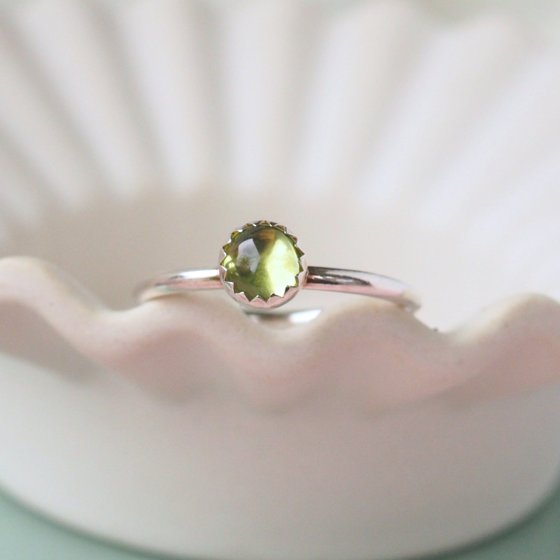 Sterling Silver and Moss Green Gemstone ring. A simple round peridot gemstone is set into Sterling Silver with a modern round band profile. The silver has a wavy zig zag edge where the stone meets the silver. The ring is shot on a white ceramic sish with a wavy edge to highlight the detail. Hand crafted in Scotland by maram jewellery