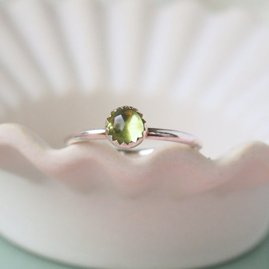 Sterling Silver and Moss Green Gemstone ring. A simple round peridot gemstone is set into Sterling Silver with a modern round band profile. The silver has a wavy zig zag edge where the stone meets the silver. The ring is shot on a white ceramic sish with a wavy edge to highlight the detail. Hand crafted in Scotland by maram jewellery