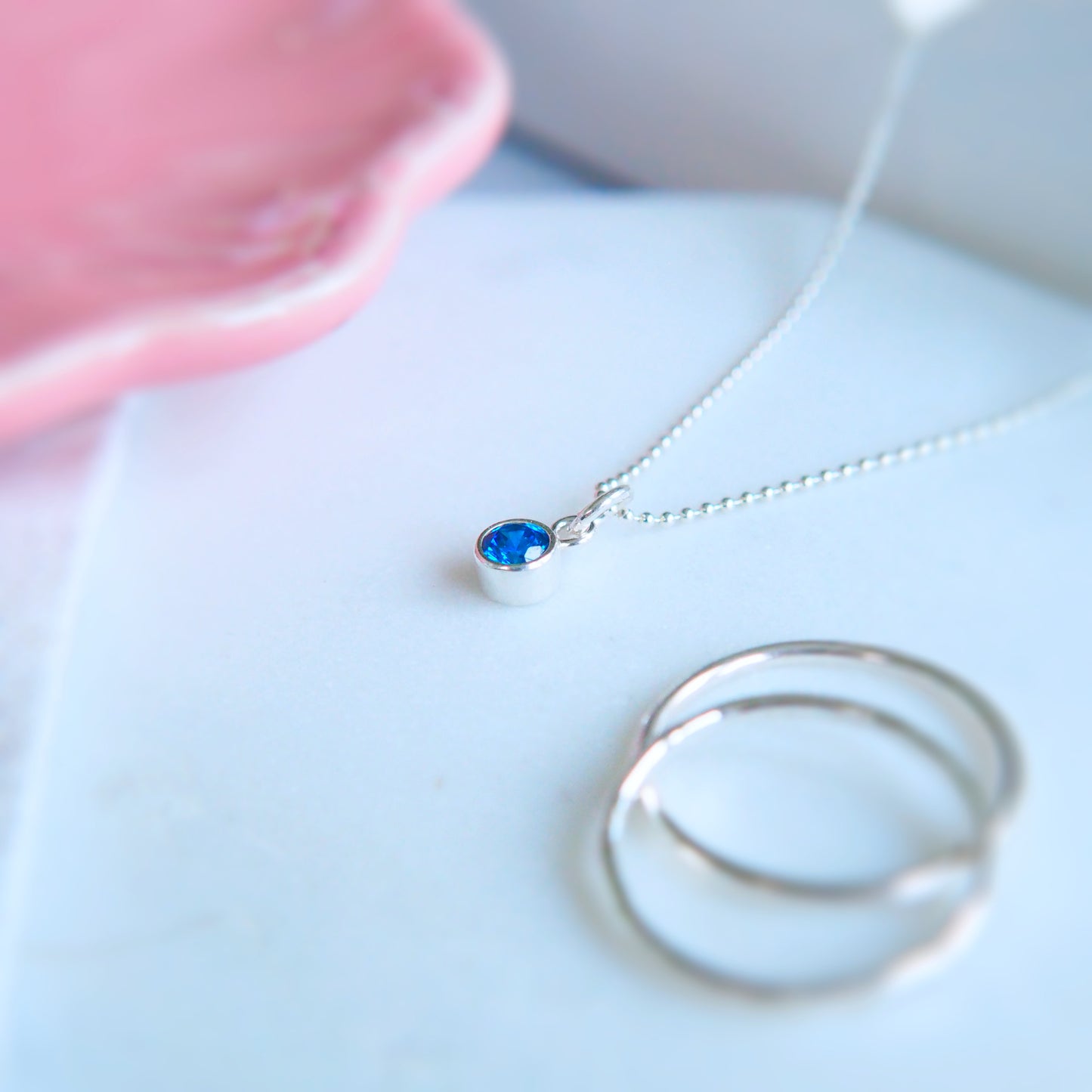 Simple Solitaire tube set Sapphire Blue Cubic Zirconia measuring 4mm set into a simple sterling silver mount on a simple silver chain with no decoration. It is pictured on a dressing table with a pink bowl, linen and a few silver rings. Made by maram jewellery in Scotland UK