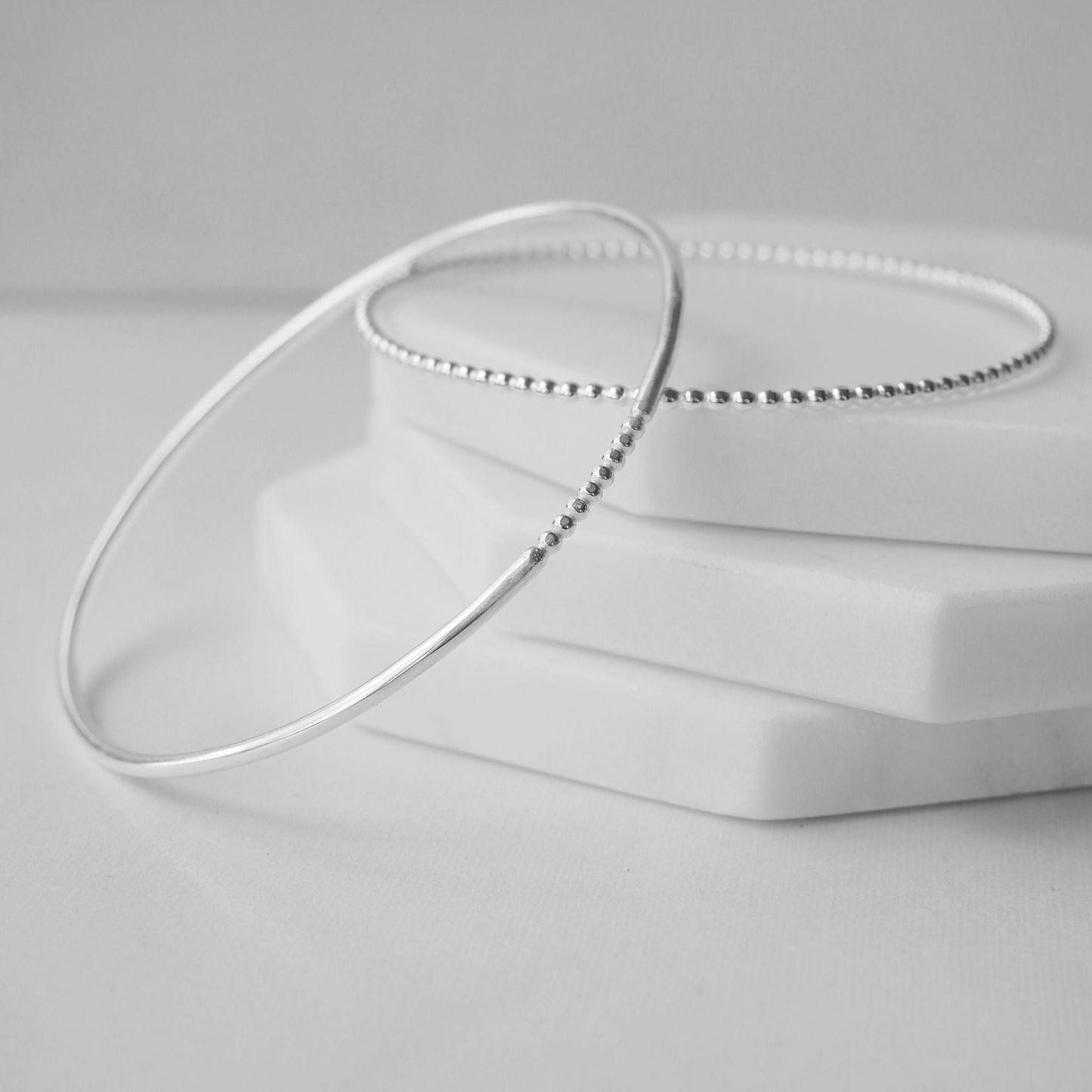 Two silver bangles on a white background, both textured. They are round in profile with a beaded texture - one with just a section and one with a full beaded looki. Handmade in Scotland by maram jewellery