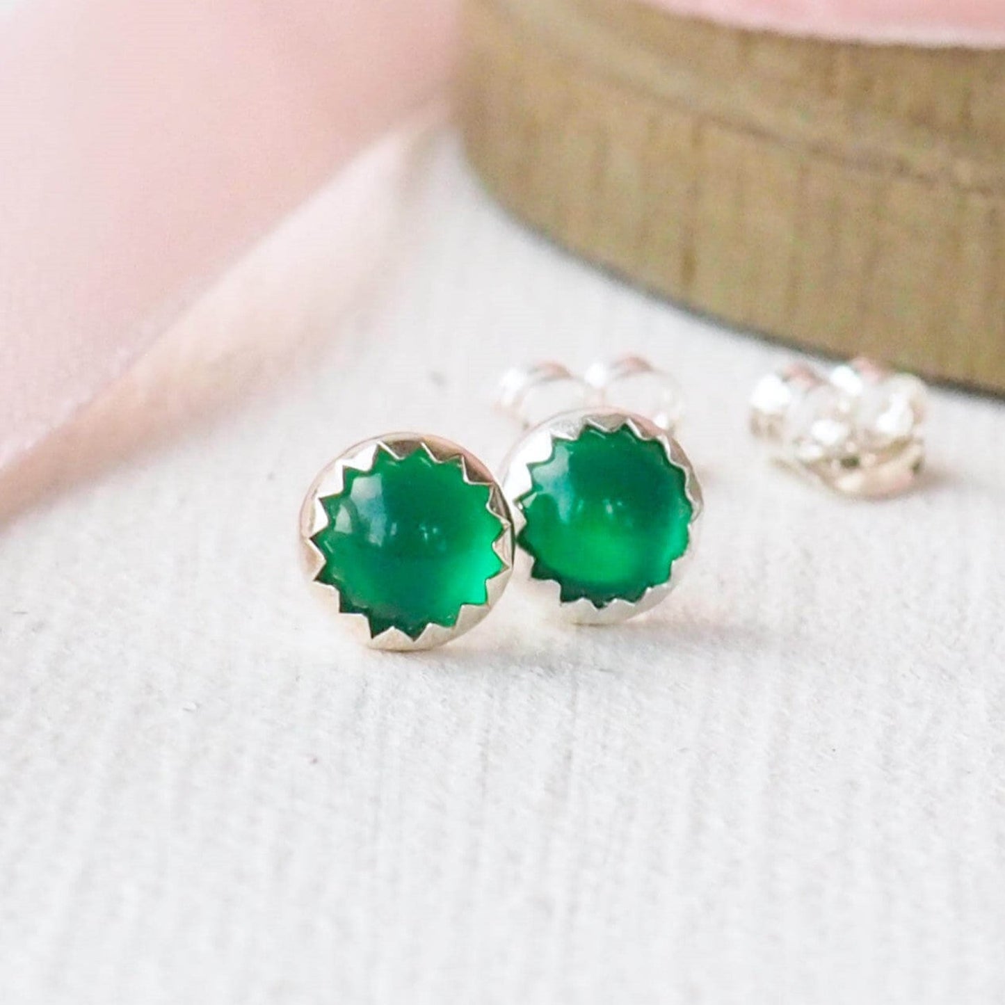 Green Agate Emerald style earrings sitting on a textured white surface with light pink ribbon reel.. The earrings are made from round cabochons measuring 5mm in green agate, birthstone for May. They are handcrafted by maram jewellery, a small indie business based in Edinburgh UK