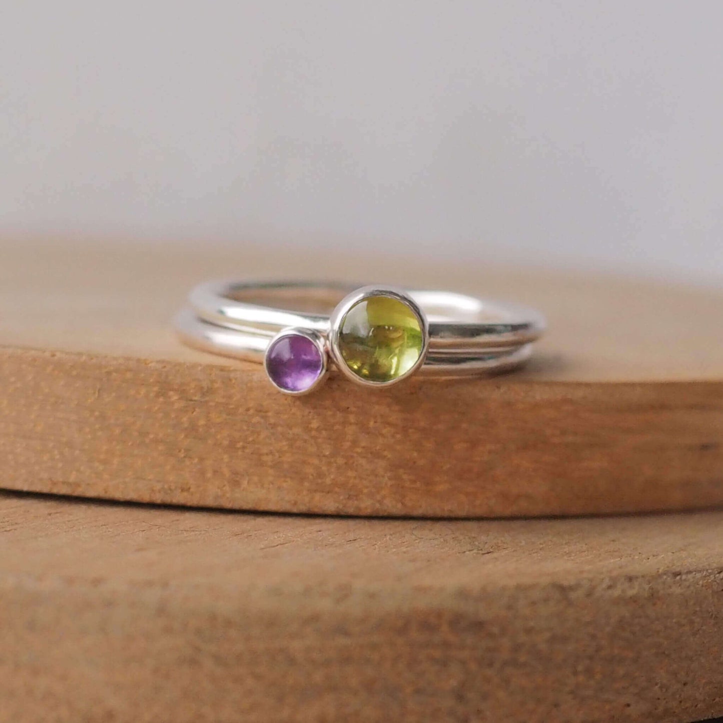 Two silver ring set with round stones. Simple round bands with a 5mm green Peridot and a 3mm purple Amethyst, birthstones for August and February. Rings are pictured on wood background. Handmade by Maram Jewellery in Scotland