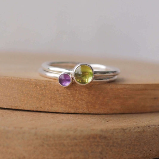 Two silver ring set with round stones. Simple round bands with a 5mm green Peridot and a 3mm purple Amethyst, birthstones for August and February. Rings are pictured on wood background. Handmade by Maram Jewellery in Scotland