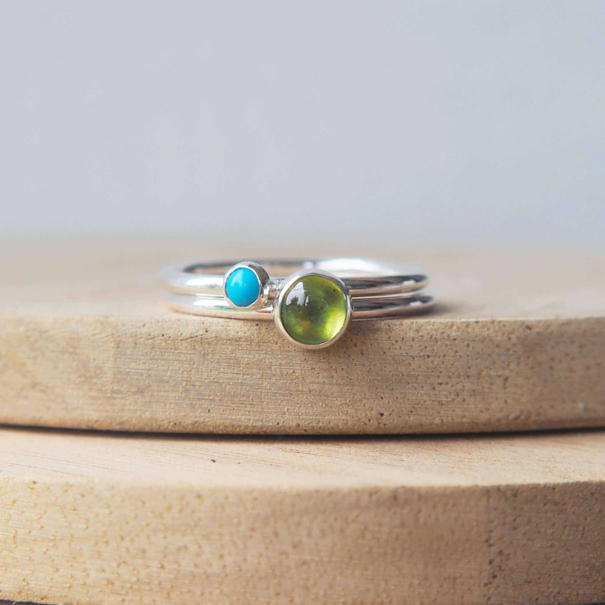 Two silver ring set with round stones. Simple round bands with a 5mm green Peridot and a 3mm turquoise, birthstones for August and December. Rings are pictured on wood background. Handmade by Maram Jewellery in Scotland