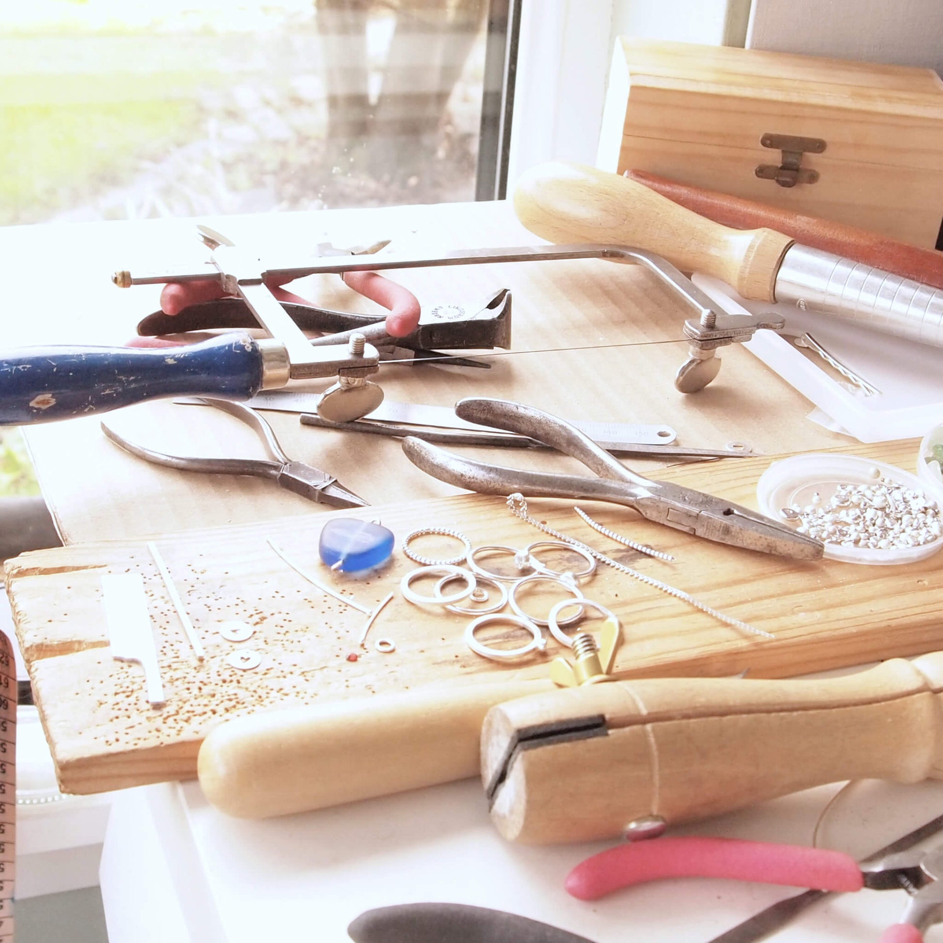 Maram Jewellery's sunny studio workbench. Image shows a light filled desk with jewellers tools, pliers and saw, alongside silver jewellery in the process of being made.