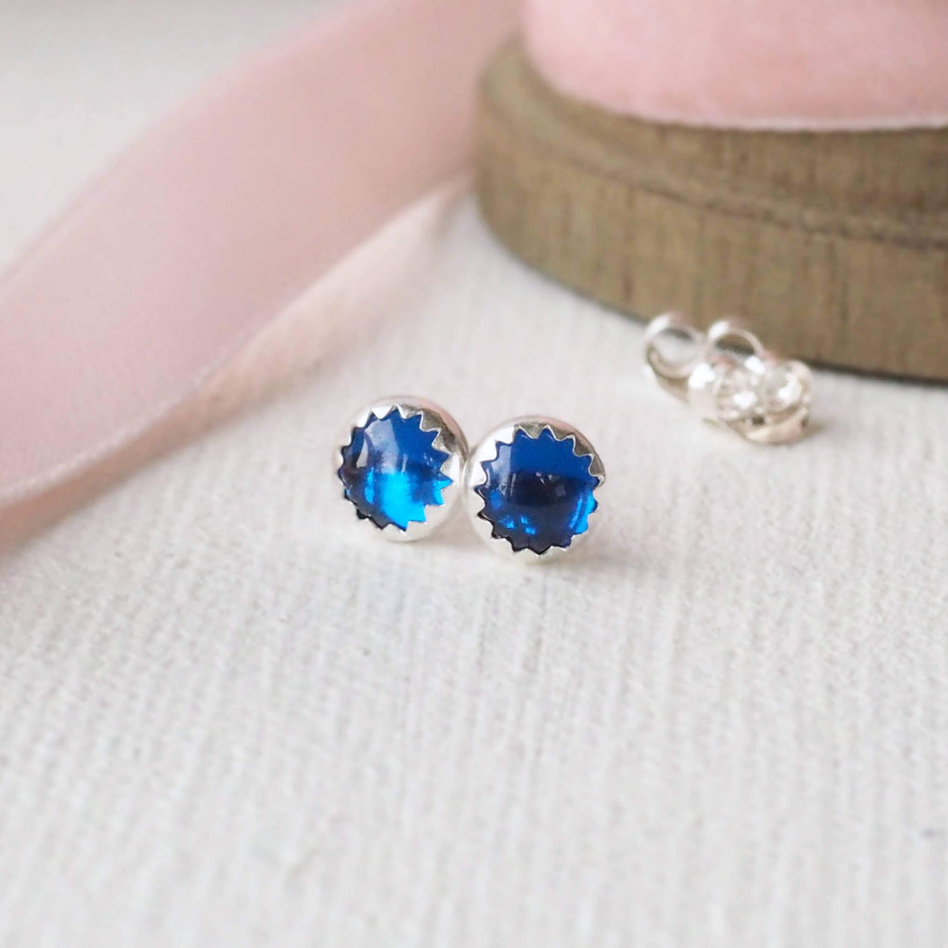 Lab Sapphire Bright Blue gemstone studs made from a pair of round 5mm cabochon cut Blue lab grown sapphires. they are set by hand into a simple sterling silver mount and are pictured on a white woven background with pale pink ribbon. Handcrafted in Scotland by maramjewellery