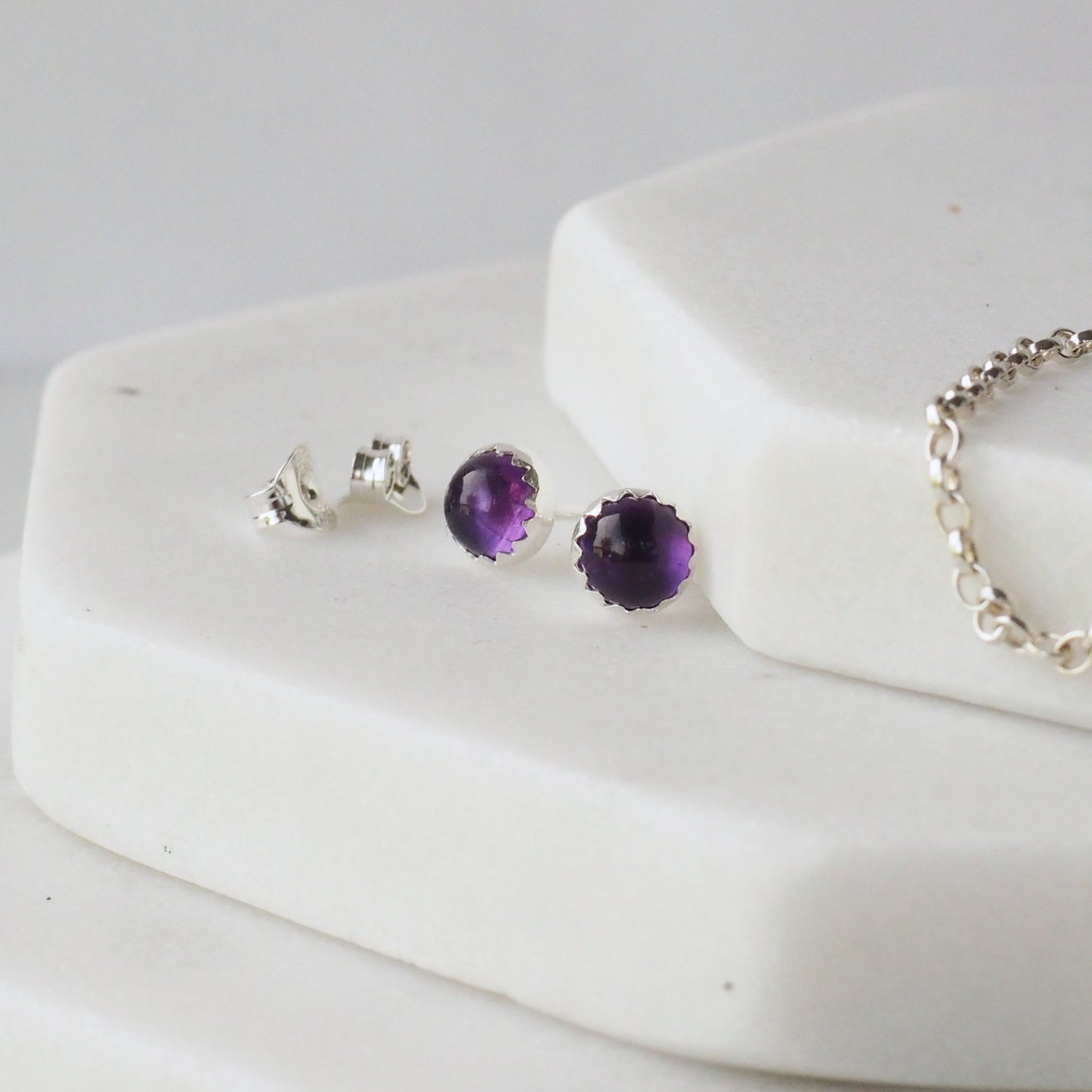 Amethyst February Birthstone Earrings in Deep Purple and Sterling Silver. The earrings are pictured on a white marble background with silver chain and earring backs alongside it. Handcrafted by maram jewellery in Edinburgh UK