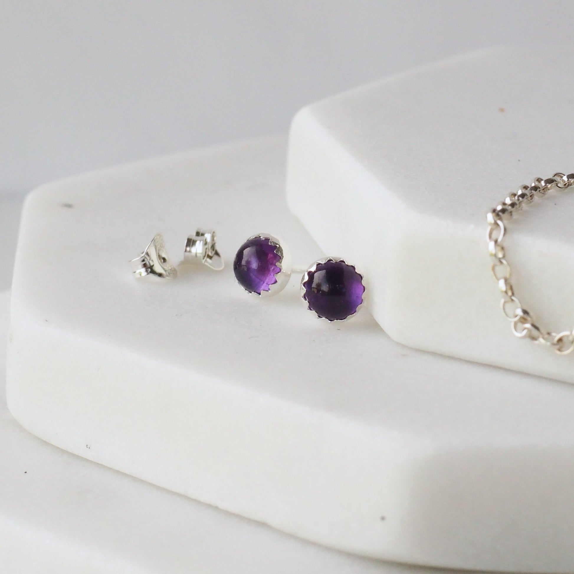 Amethyst February Birthstone Earrings in Deep Purple and Sterling Silver. The earrings are pictured on a white marble background with silver chain and earring backs alongside it. Handcrafted by maram jewellery in Edinburgh UK