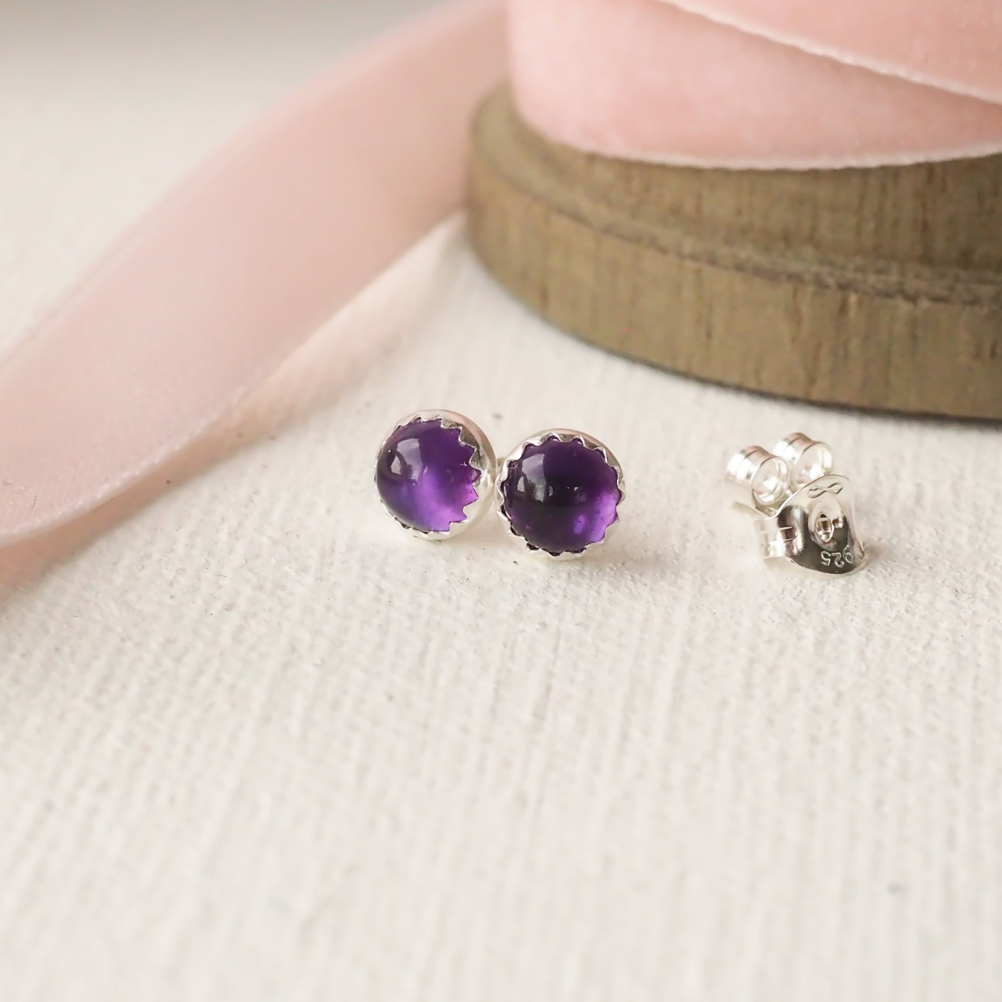 Purple Amethyst round stud earrings pictured on a white linen texture with pink ribbon in the background. The earrings measure 5mm and are round, Sterling Silver and deep purple in colour./ Handcrafted by maram jewellery in Edinburgh UK
