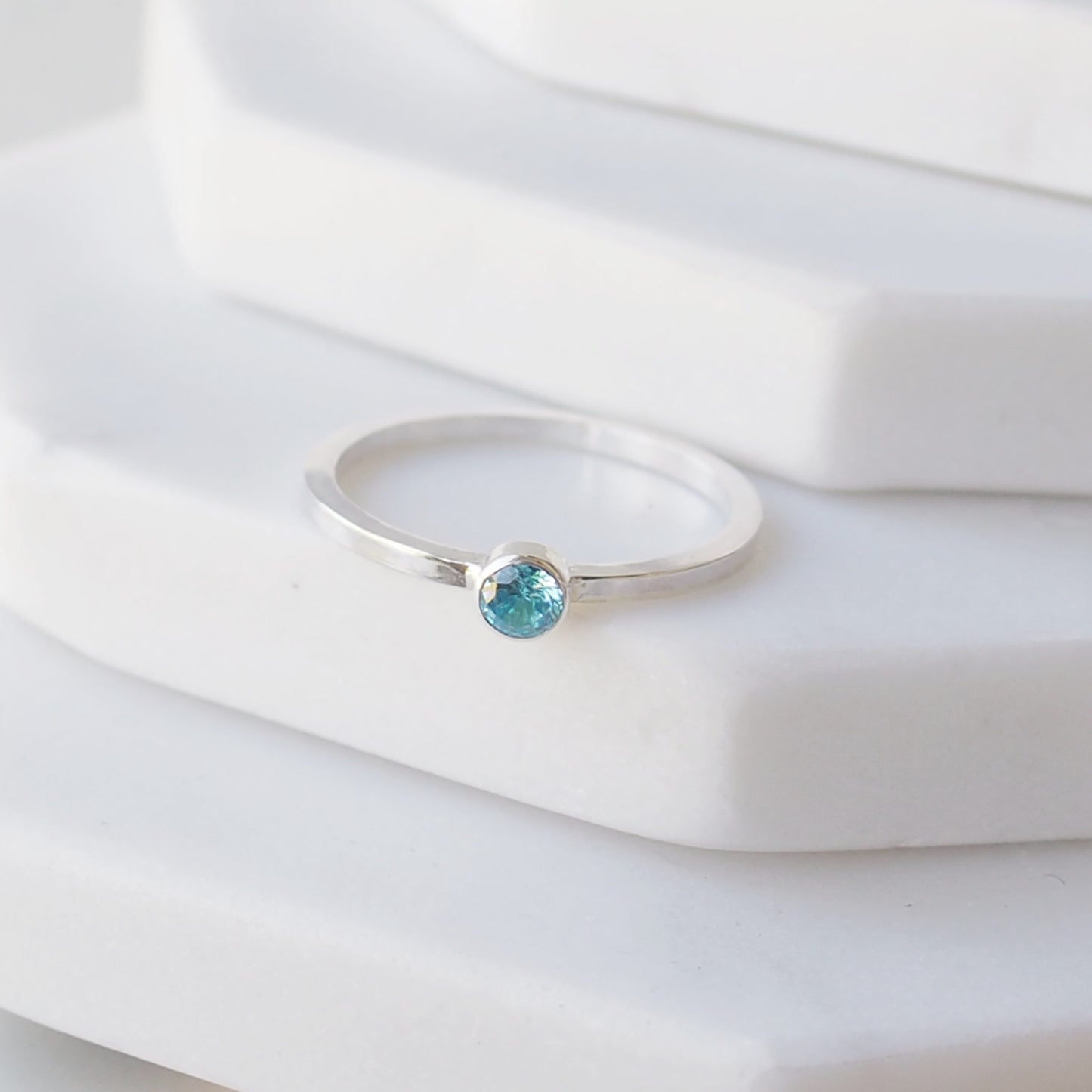 Silver ring with an aquamarine coloured gemstone. The ring is simple in style with no embellishment , with a square wire band 1.5mm thick with a simple aquamarine 4mm round cubic zirconia stone set in an enclosed silver setting. The ring is Sterling Silver and made to your ring size. Handmade in Scotland by Maram Jewellery