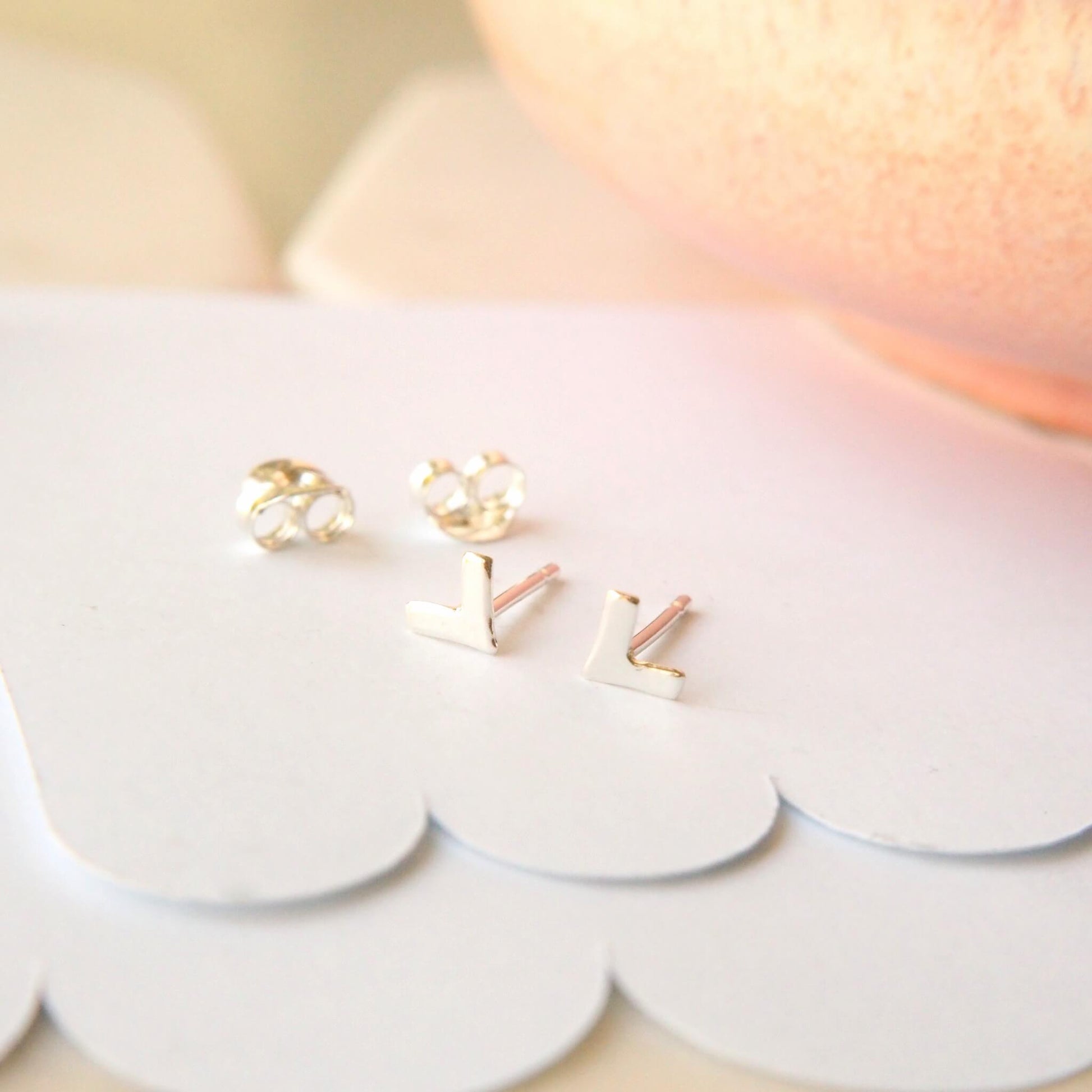 Small silver stud earrings in a chevron shape, the earrings measure 6mm in height. the studs are handmade by maram jewellery and are pictured on a scallop card background with a pale pink dish in the background