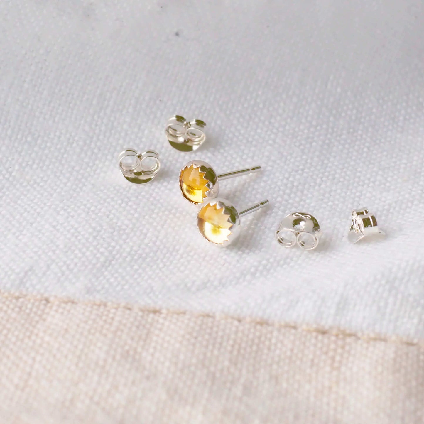 Beautifully simple modern gemstone stud earrings with a warm yellow round citrine set by hand into a modern minimalist sterling silver surround, sitting with earring backs on a linen cloth.The earrings are 5mm in size, round and come with butterfly backs.Citrine is the birthstone for November. These stud earrings are handcrafted by a small business in Scotland UK by designer jeweller Maram Jewellery