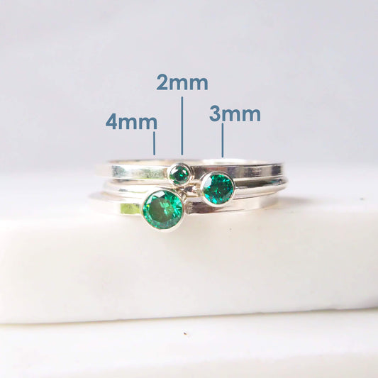 Trio of Emerald Green Silver Cubic Zirconia Solitaire rings with round stones. Each of the rings have a different sized gem in a 2mm, 3mm and 4mm size with sizes marked in text.. Pictured on a simple white marble background. Rings handmade by maram jewellery in Scotland UK