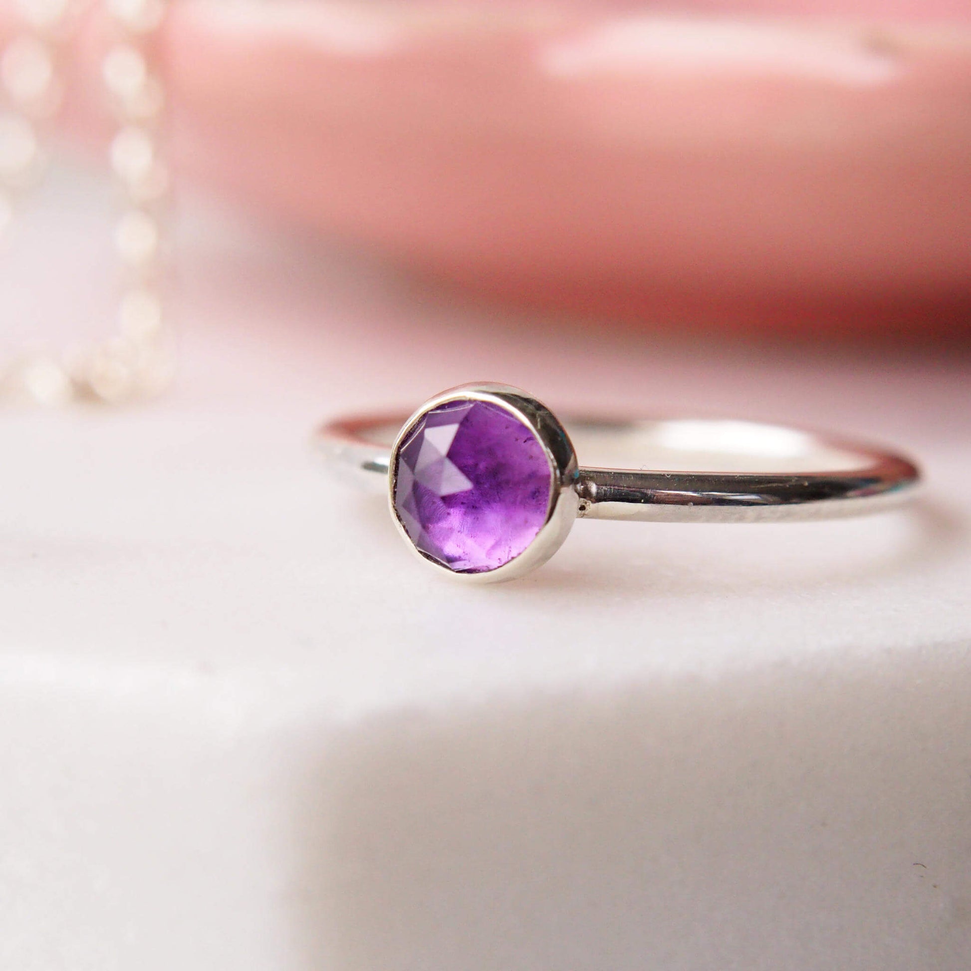 Facet Amethyst 5mm round ring on a white marble table with a pink trinket dish behind. Hand crafted sterling silver February birthstone ring by maram jewellery