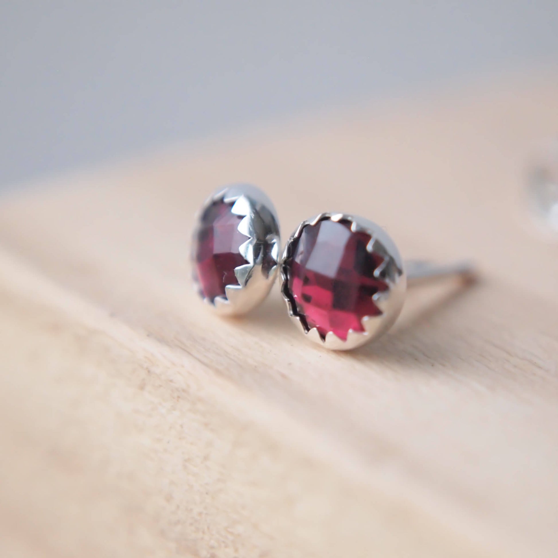 A pair of modern style gemstone earrings in sterling silver and garnet. round gemstones with a face cut measuring 5mm, set simple into a modern silver surround with a serrated edge that is very subtle. Hand crafted in a small jeweller studio in Scotland UK