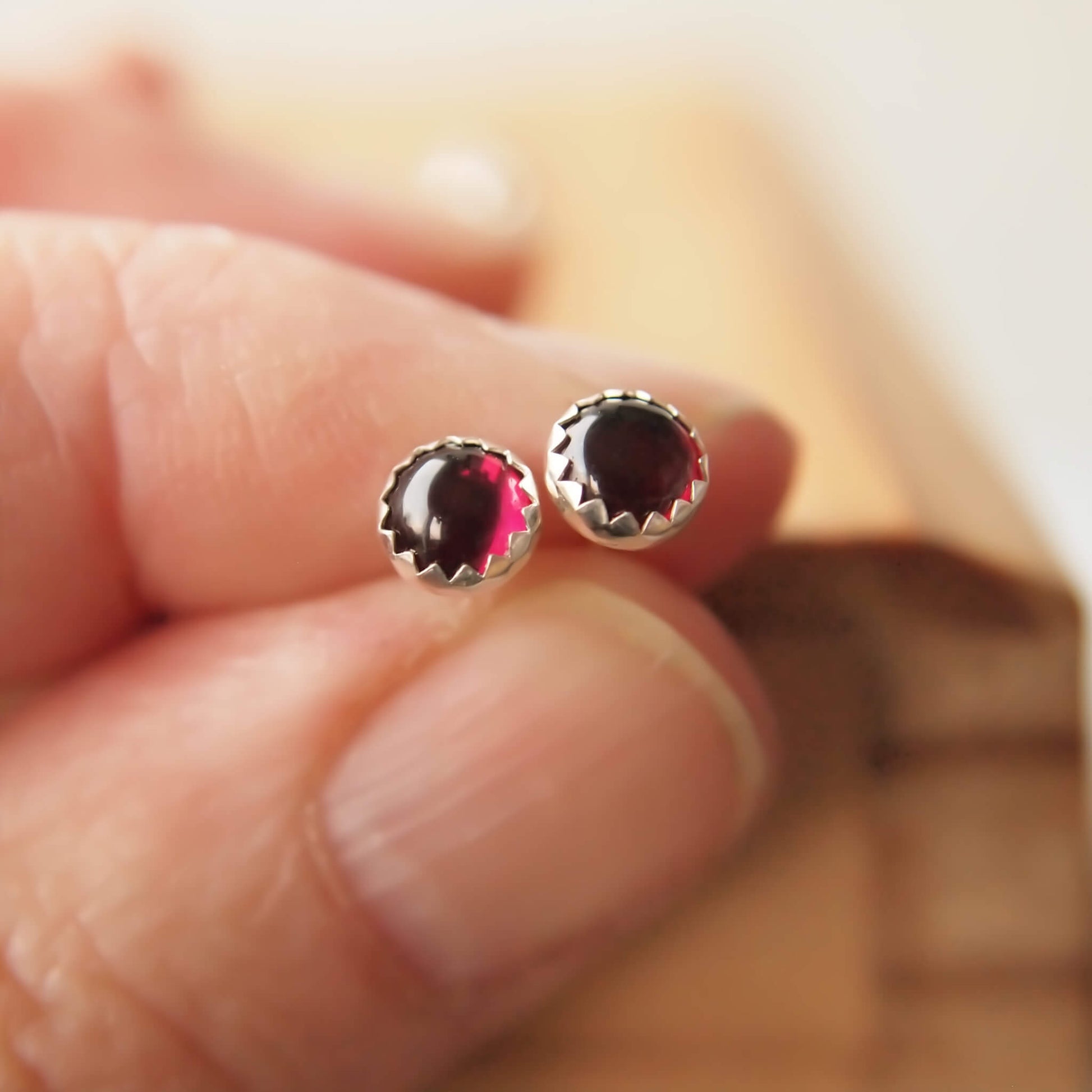 Small Gemstone studs made by maram jewellery in Scotland UK. The earrings are handcrafted from Sterling Silver, simple in design and have a matching pair of 5mm round garnets, birthstone for January. The earrings are minimalist and modern in design and are shown held in a hand to show scale