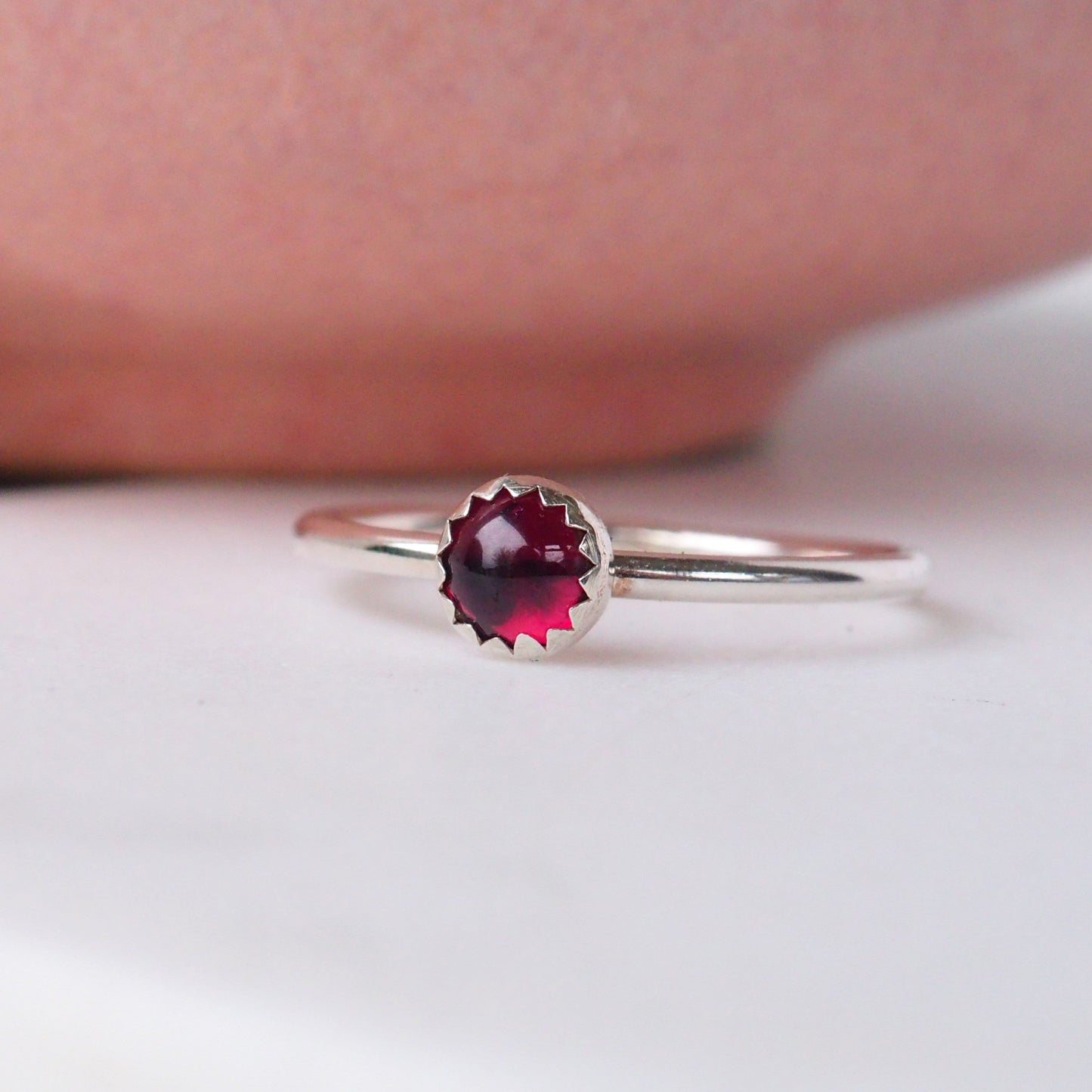 Modern style simple solitaire ring with a round Garnet Gemstone. The ring has a fully round band made to your size, with a 5mm round gemstone set carefully into the silver with an unusual serrated edge where the gem meets the silver. It is hand crafted by maram jewellery in Scotland and photographed on a white background with an out of focus pink bowl just showing.