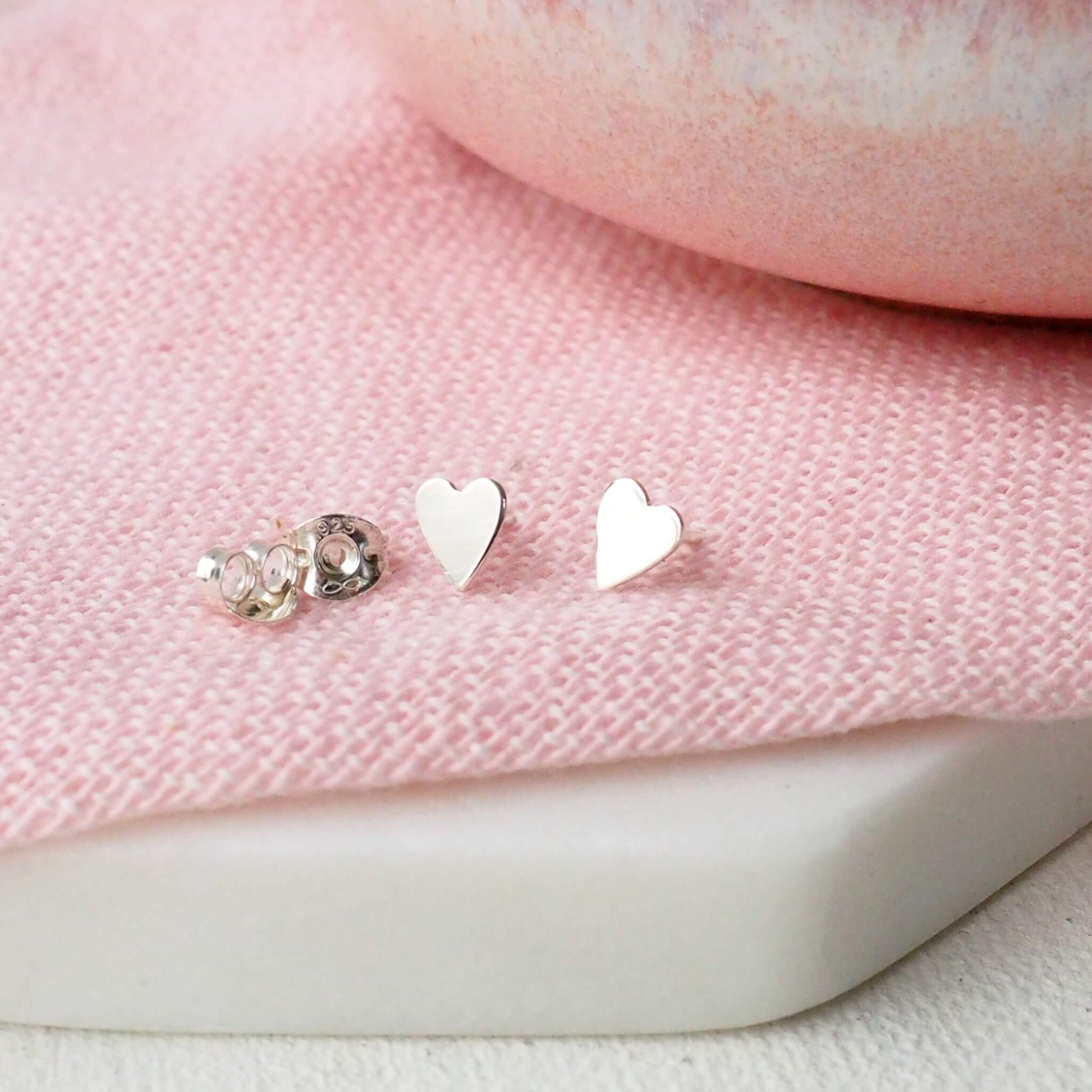 Tiny silver heart shaped studs pictured on a pink linen cloth with a pink bowl in the background. The earrings are Sterling Silver around 5mm in size and are handmade by maram jewellery