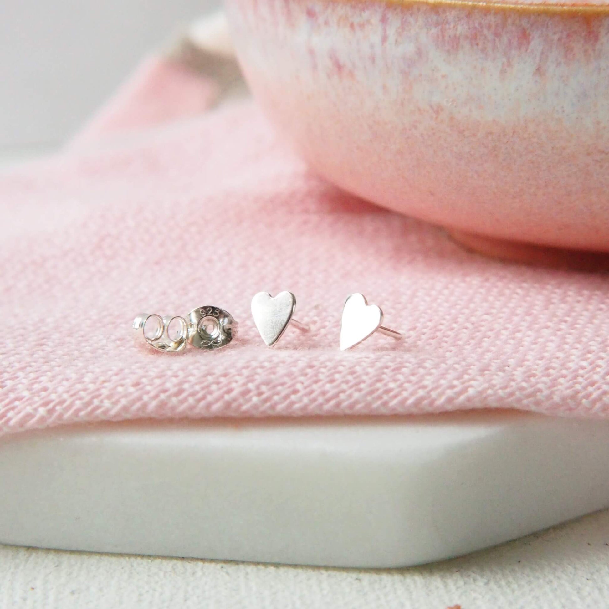 Tiny silver heart shaped studs measuing 5mm in size pictured on a pink linen cloth with a pink bowl in the background. The earrings are handmade by maram jewellery