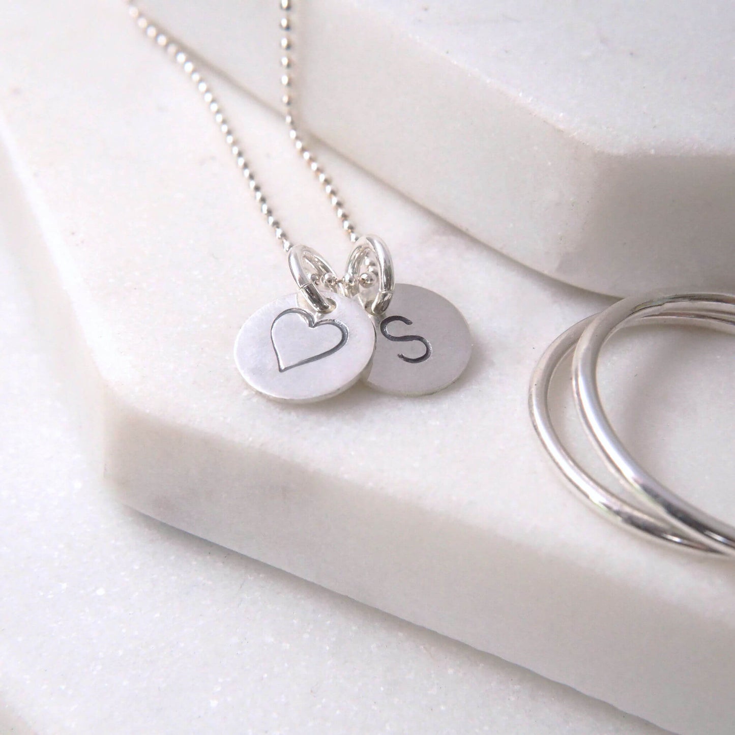 Silver letter and heart token charms on a simple silver chain. Two small silver discs, one with a heart and the other with an initial S though you can choose a different one too. Subtle and understated necklace handmade by maram jewellery in UK