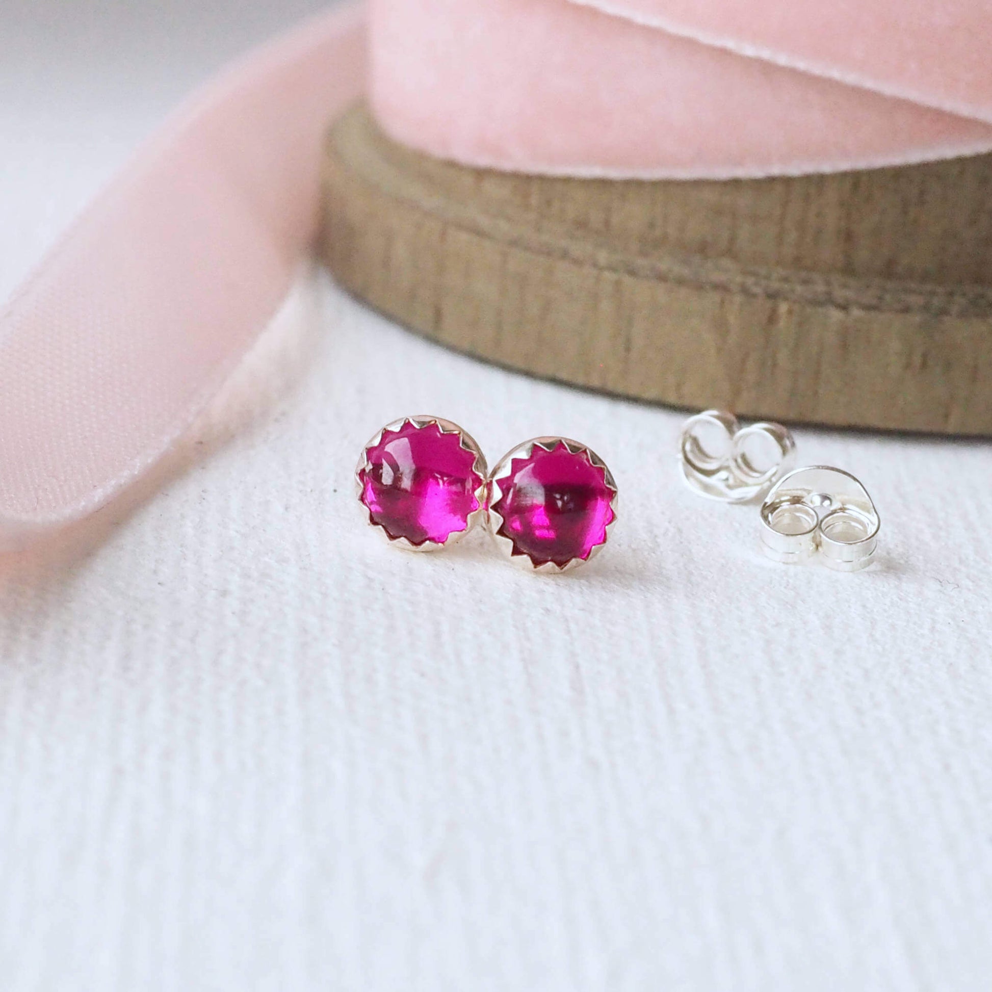 Lab Ruby Pink gemstone studs made from a pair of round 5mm cabochon cut pink lab grown rubies. they are set by hand into a simple sterling silver mount and are pictured on a white woven background with pale pink ribbon. handcrafted in Scotland by maramjewellery