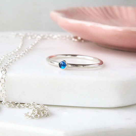 Blue Sapphire small gemstone ring in Sterling Silver. The ring has a round modern band and a simple 3mm round gemstone in Lab Sapphire.  Pictured on a white marble with a pink bowl in the background. Handcrafted by maram jewellery in Edinburgh UK