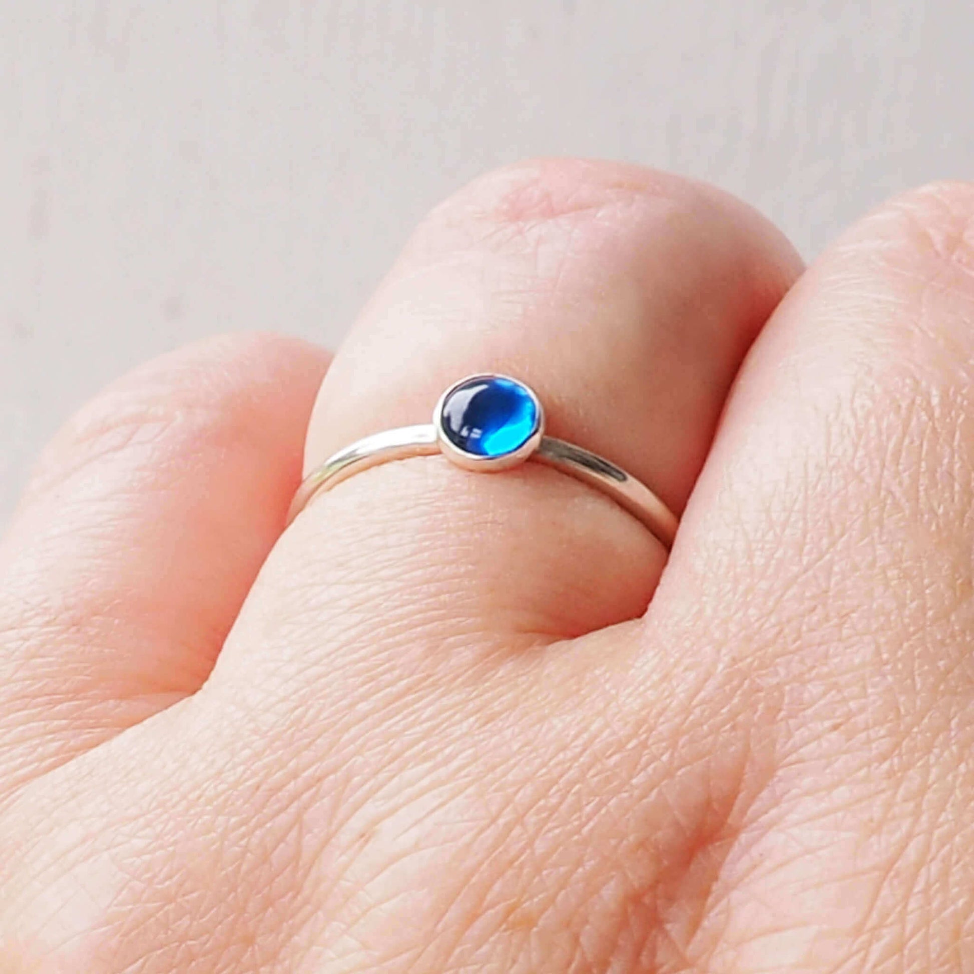 Silver Ring with a round gemstone in a simple style. The gem is a 5mm round lab sapphire cabochon in a bright primary blue. It's worn on a hand to show scale. Handmade by maram jewellery in Scotland