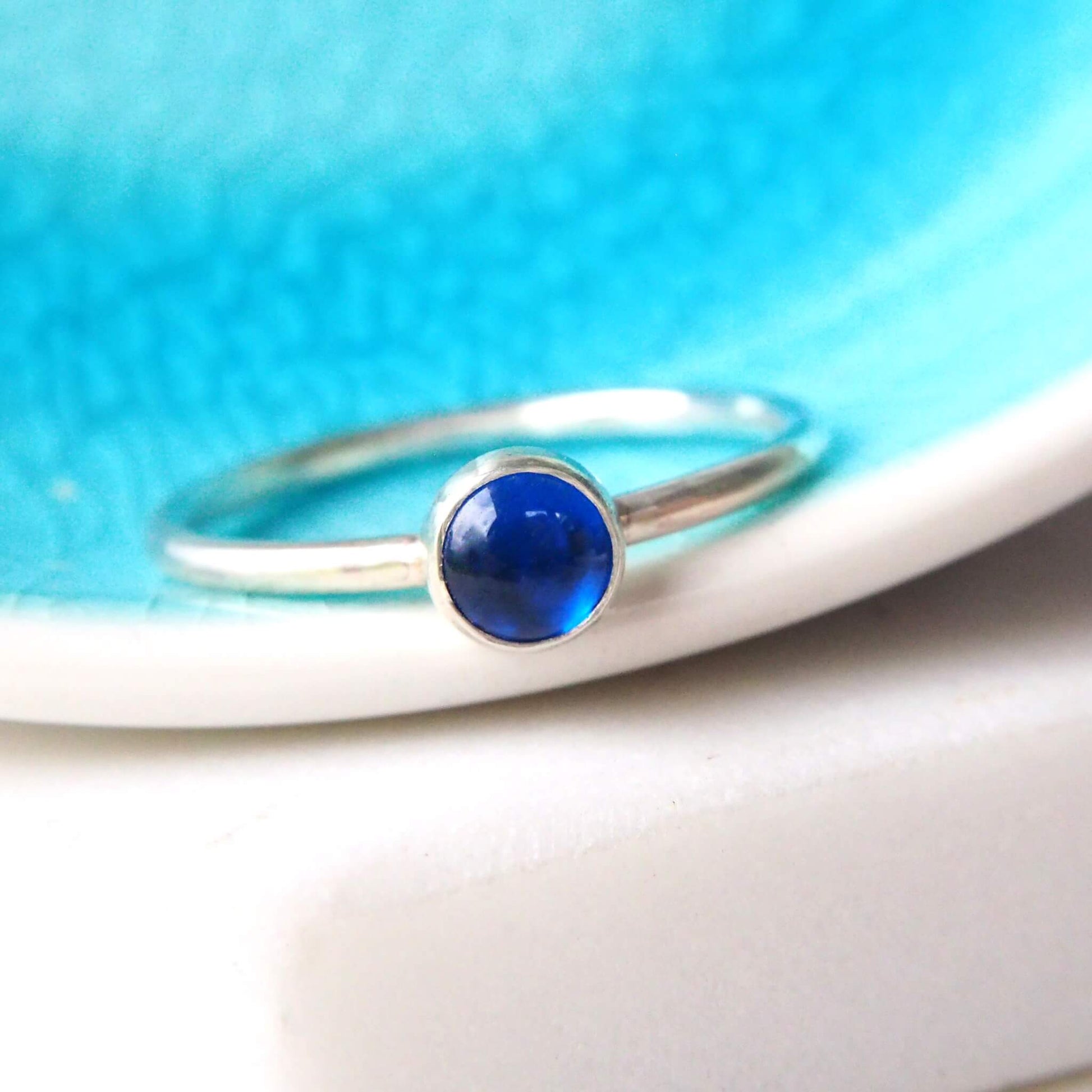 Modern Sapphire solitaire ring pictured on a bright blue ceramic dish. The ring in made in Sterling Silver with a Lab created Sapphire, round in shape in a 5mm size. Made in a modern simple minimalist style by maram jewellery in Edinburgh Scotland