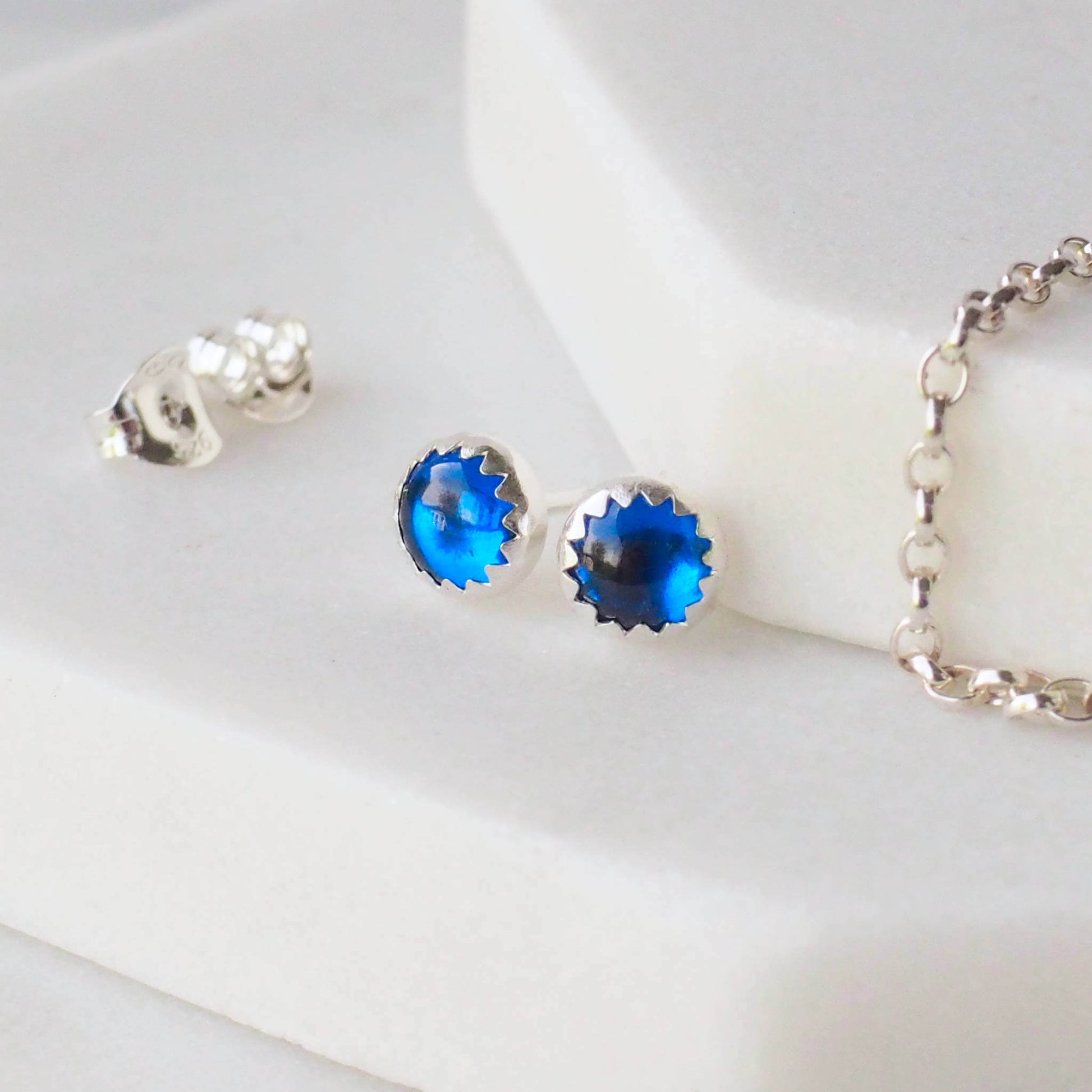 Lab Sapphire Earrings sitting on a hexagonal marble surface. The earrings are made from round cabochons measuring 5mm in a bright blue lab created sapphire, birthstone for September. They are handcrafted by maram jewellery, a small indie business based in Edinburgh UK