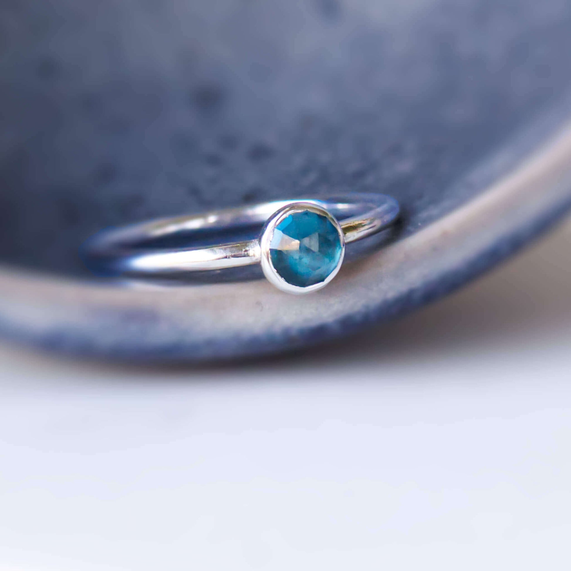 London Blue Topaz round gemstone ring. Silver ring in a minimalist style with a simple round band and a undecorated 5mm round facet cut teal coloured Blue Topaz. Petrol blue gemstone on silver pictured on a textured grey ceramic dish
