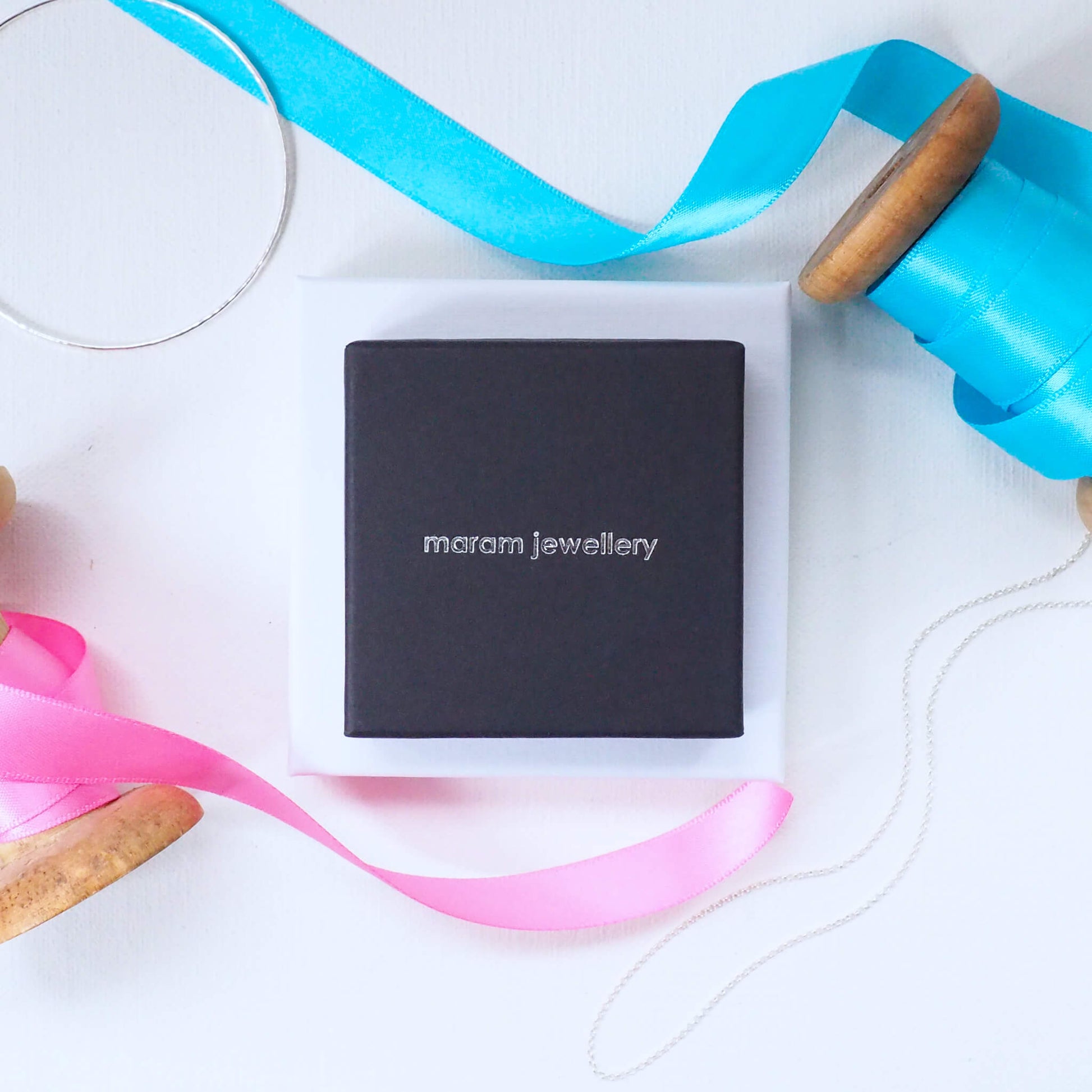 maram jewellery pendant or bangle box in black with 'maram jewellery' text in silver in the middle. shown surrounded by ribbon bangle and chain. maram jewellery is based in Scotland UK