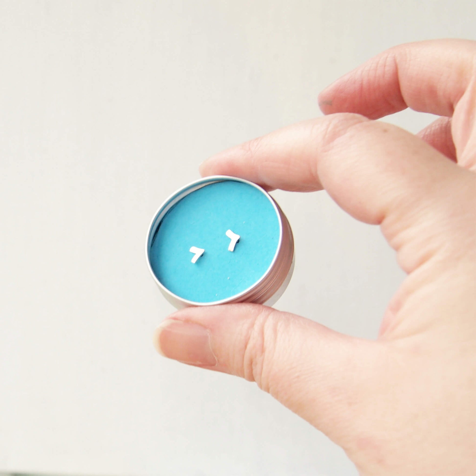 A pair of tiny stud earrings in a round aluminium tin with a bright blue insert. a hand is holding the tin between two fingers. The tin contains a pair of silver chevron earrings handmade by maram jewellery
