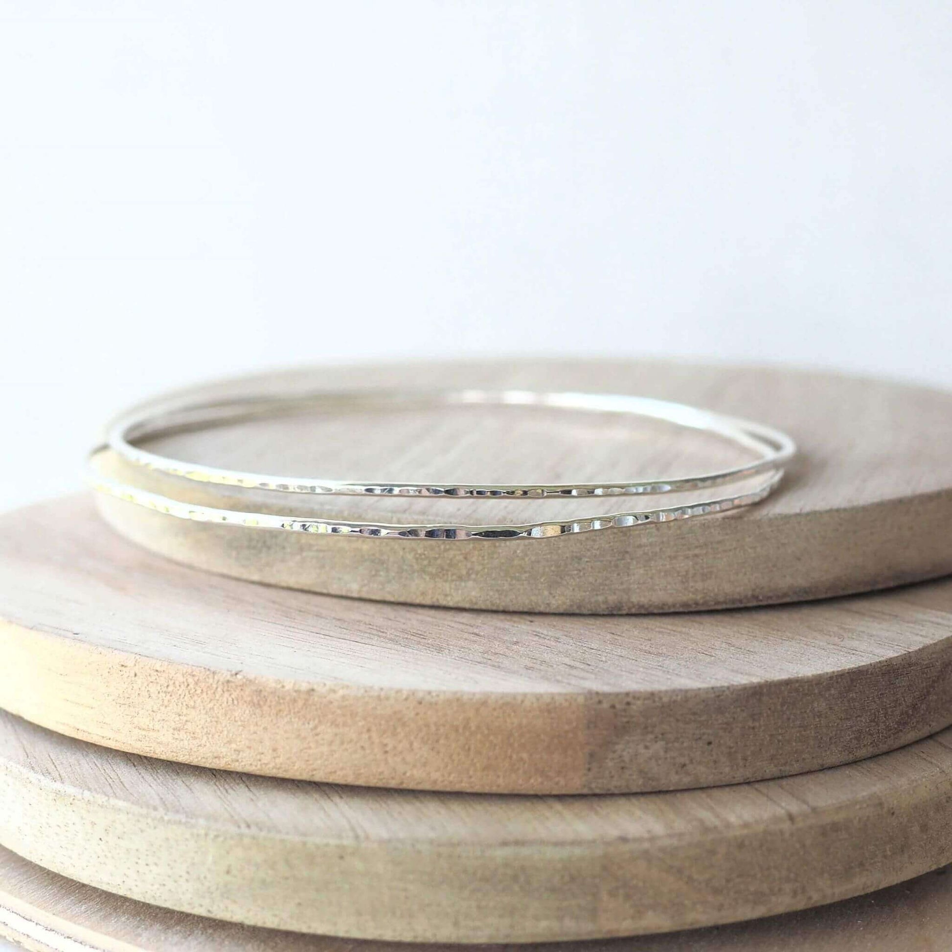 Pair of narrow hammered silver bangles designed for layering and wearing in sets. Set of two pictured on a wood background. Handmade by maram jewellery in Edinburgh UK