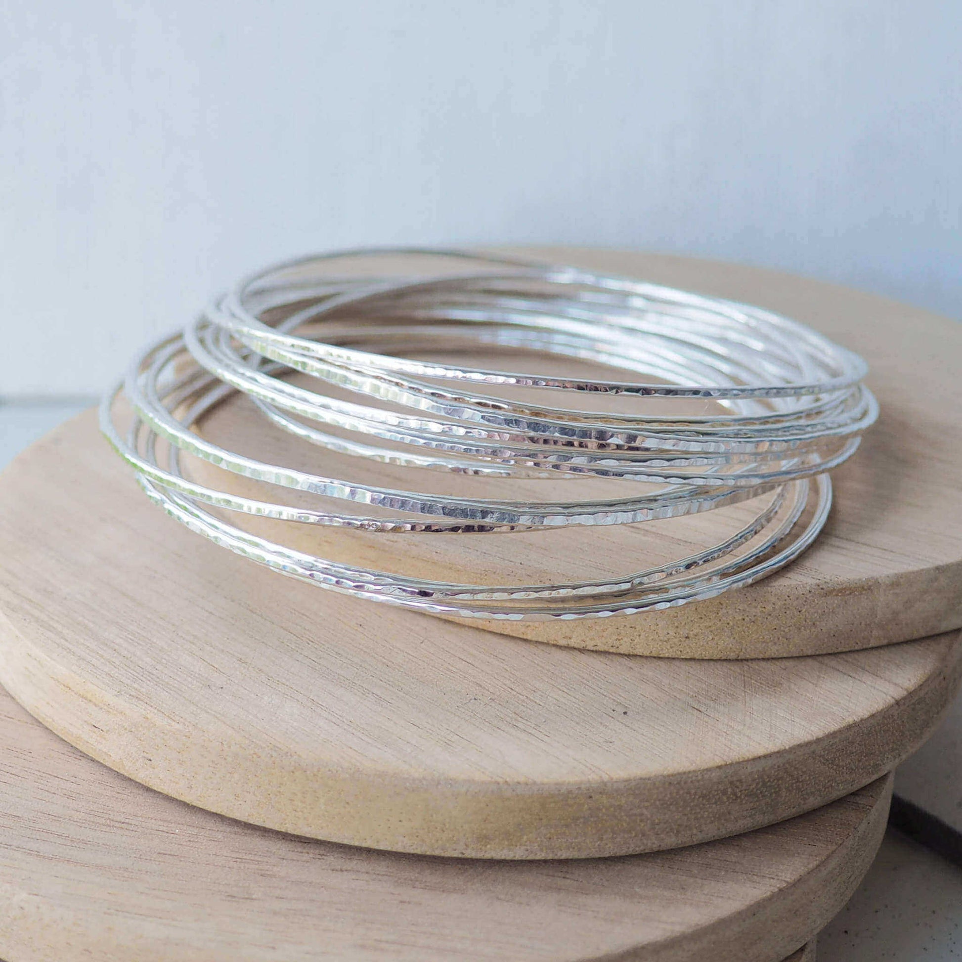 Pile of sterling silver hammered thin bangles in a stack, shimmering against a pale wood background. bangles are handmade in a choice of sizes by maram jewellery in Scotland UK