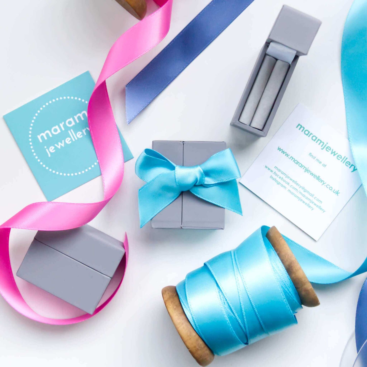maram jewellery ring packaging - a light grey box wrapped in colourful ribbon. Image shows box open and closed with a ribbon tie  with a selection of pink turquoise and blue ribbons in the background. maram jewellery is a small boutique jeweller in Scotland UK