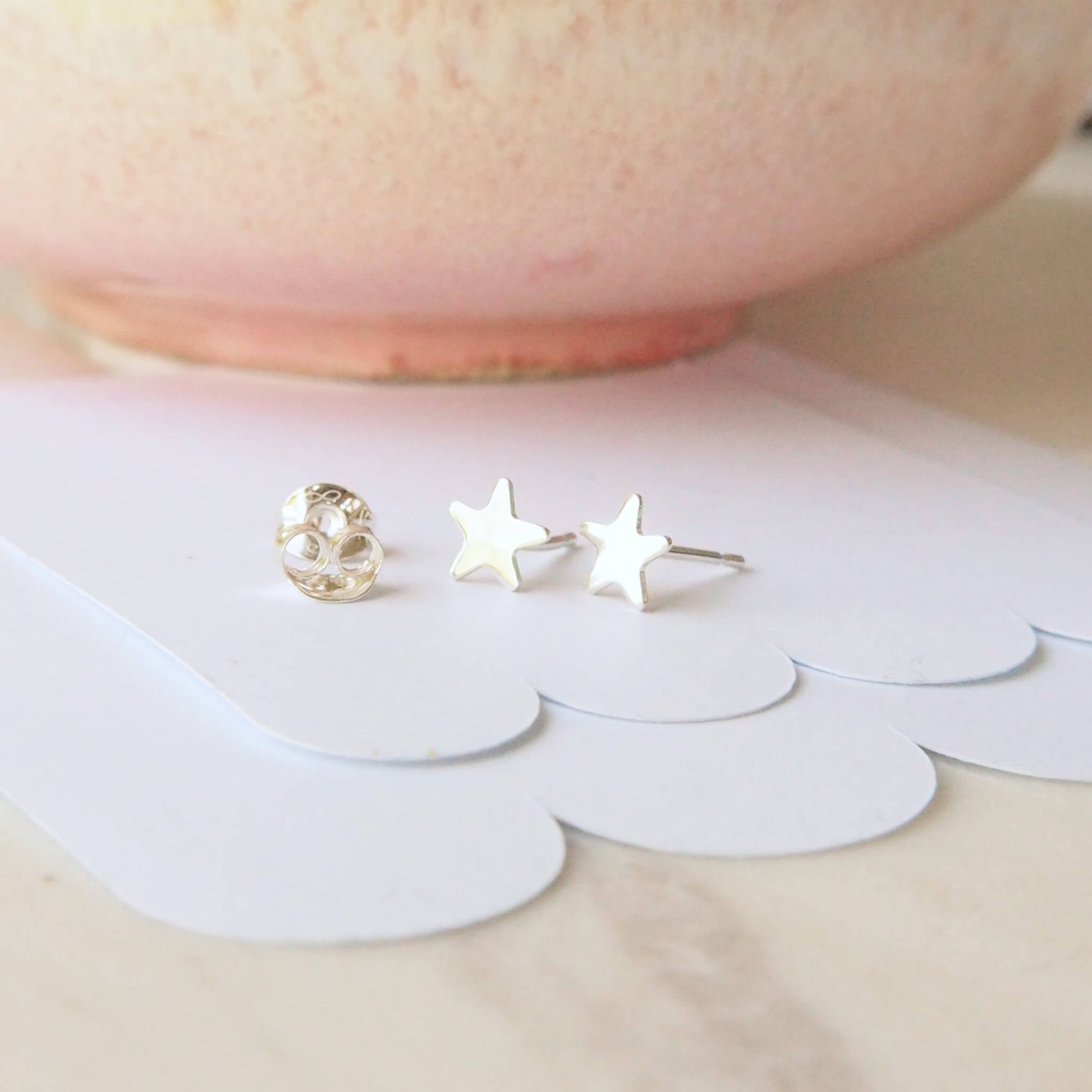 Simple small sterling silver star earrings pictured on a white scallop card background with a pink bowl in the background. The earrings measure 5mm in size and are handmade by maram jewellery in Edinburgh UK